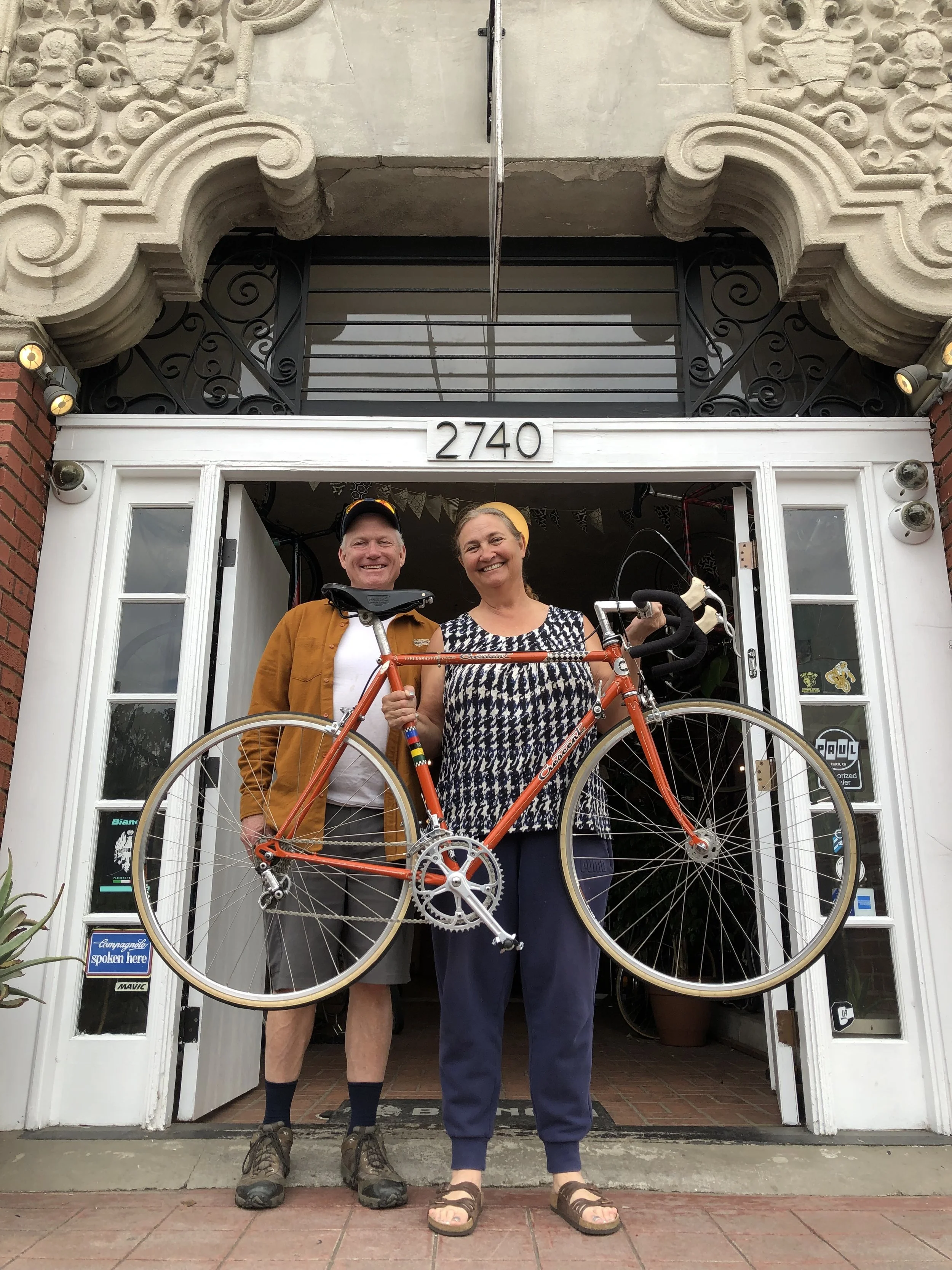 Smiling man and woman standing in front of building with open door, woman holding bicycle frame, building address 2740 above door.