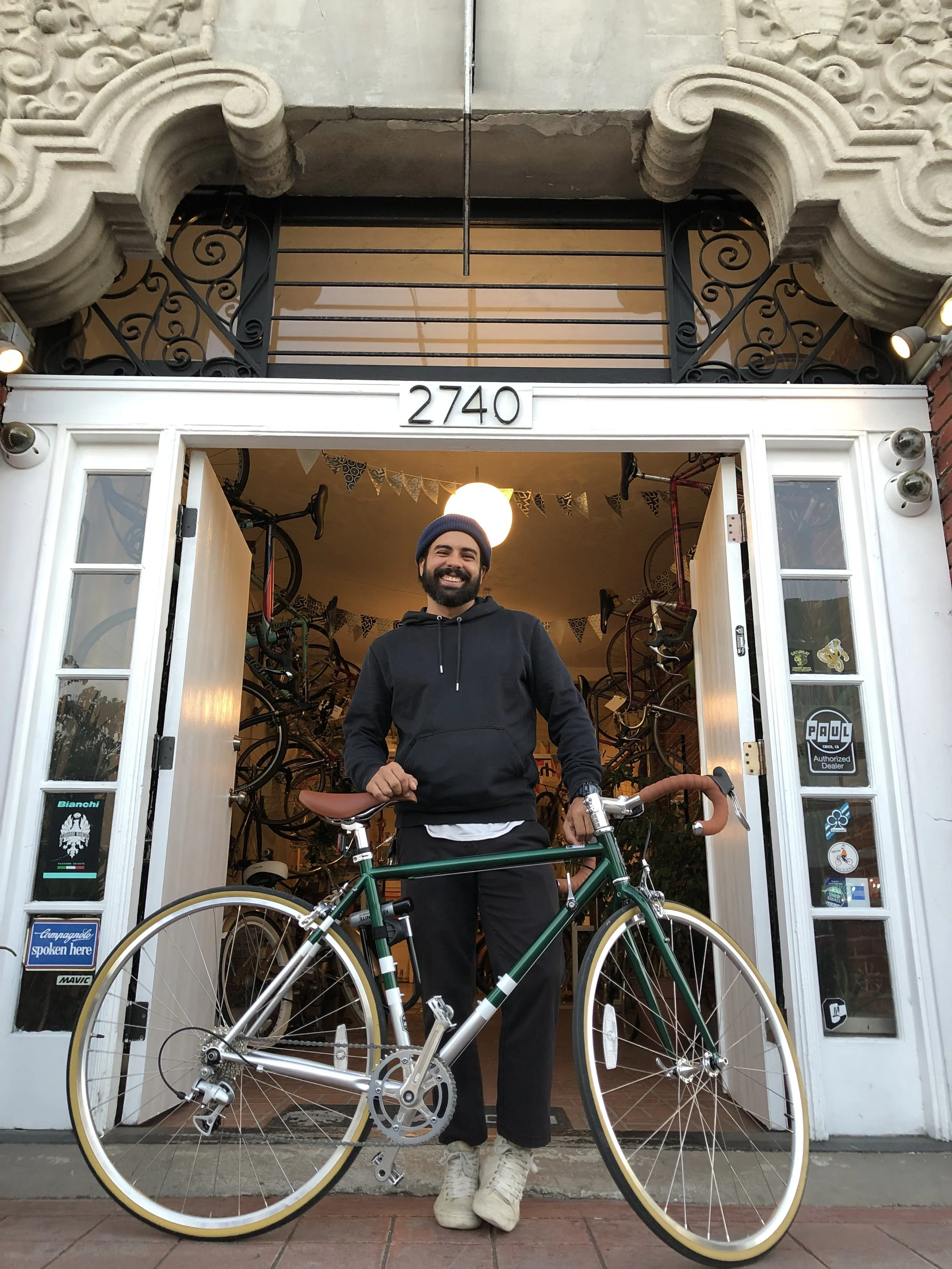 A man standing with a bicycle inside a bike shop, smiling at the camera, with bikes hanging on the wall behind him.