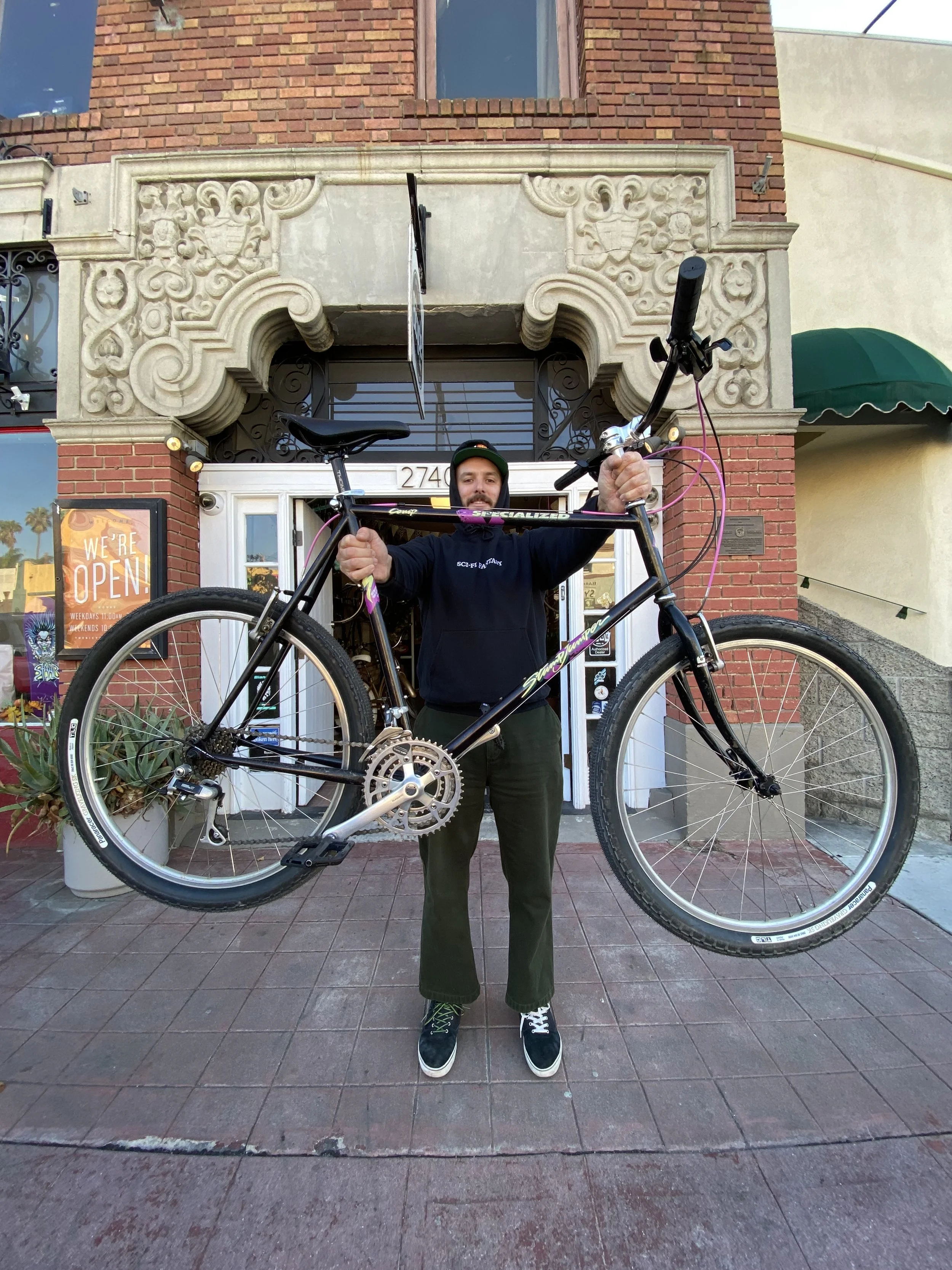 A man holding a large bicycle above his head in front of a building with a decorative stone archway and a sign that reads 'We're Open'.