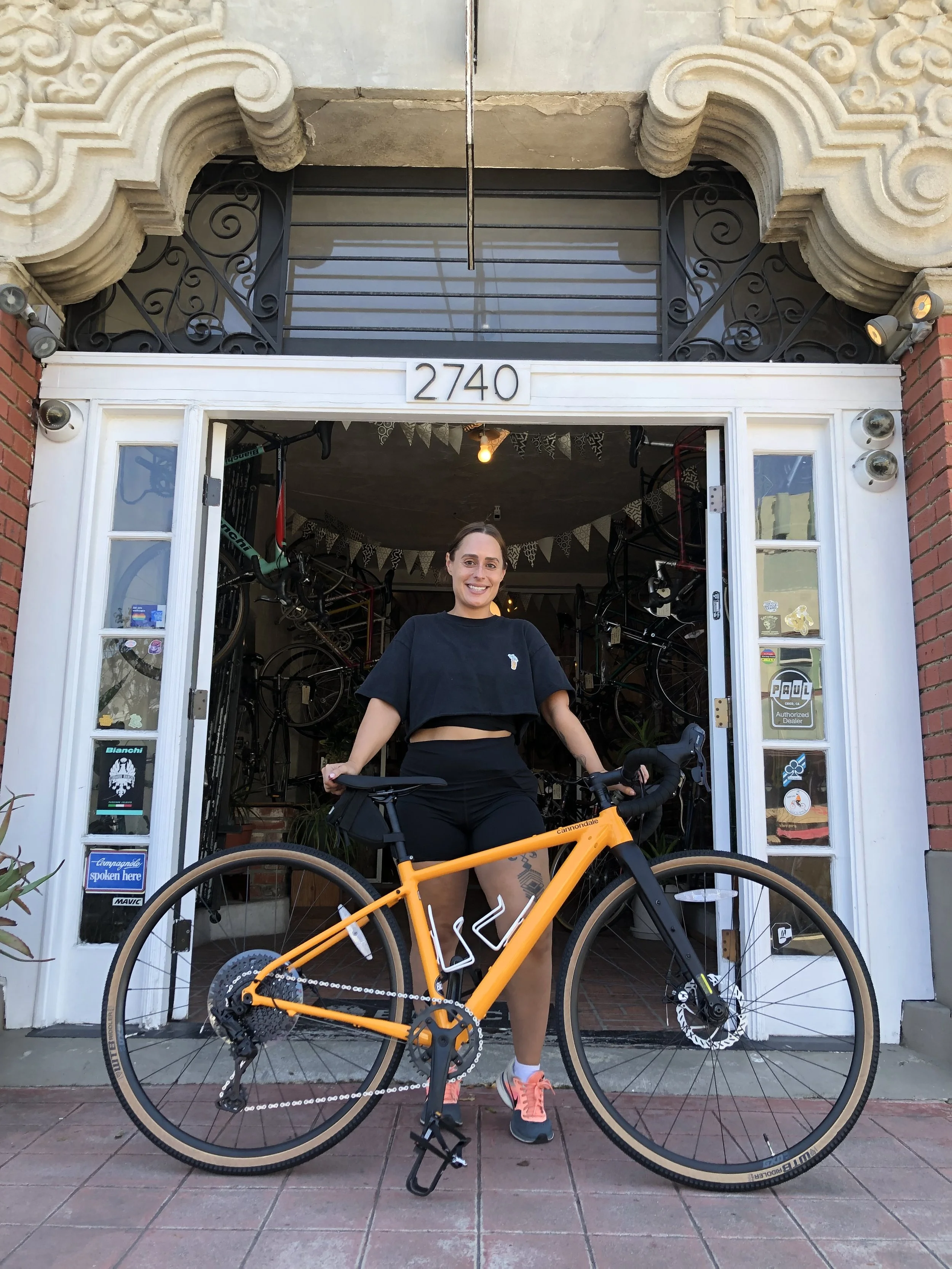 A woman standing in front of a bicycle shop with a bright orange bicycle, wearing black shorts and a black crop top, smiling at the camera.