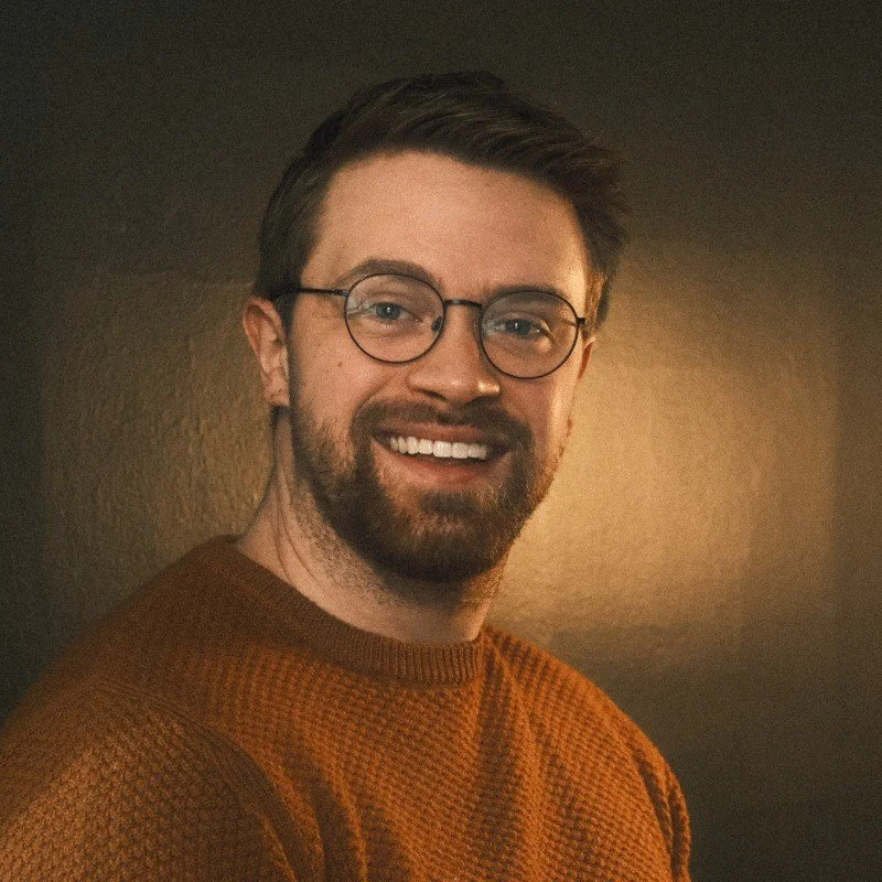 A smiling man with glasses and a beard, wearing a brown sweater, standing against a dark background.