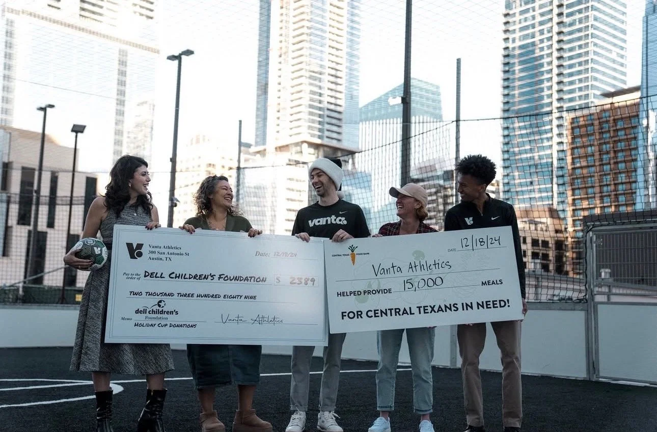 Group of five people standing on a sports field in an urban area, holding large checks and smiling.