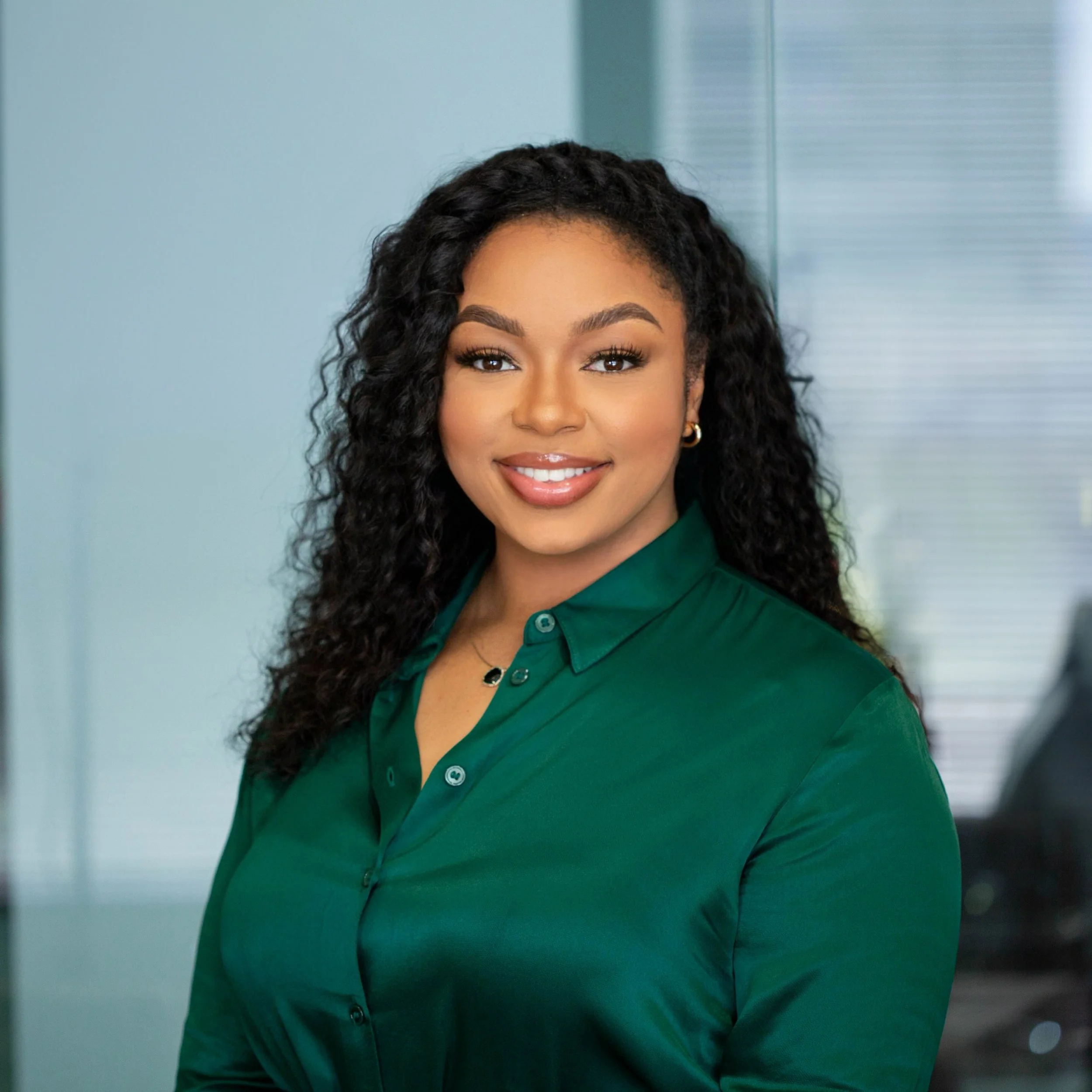 A woman with long, curly black hair wearing a green button-up shirt, smiling in an office setting.