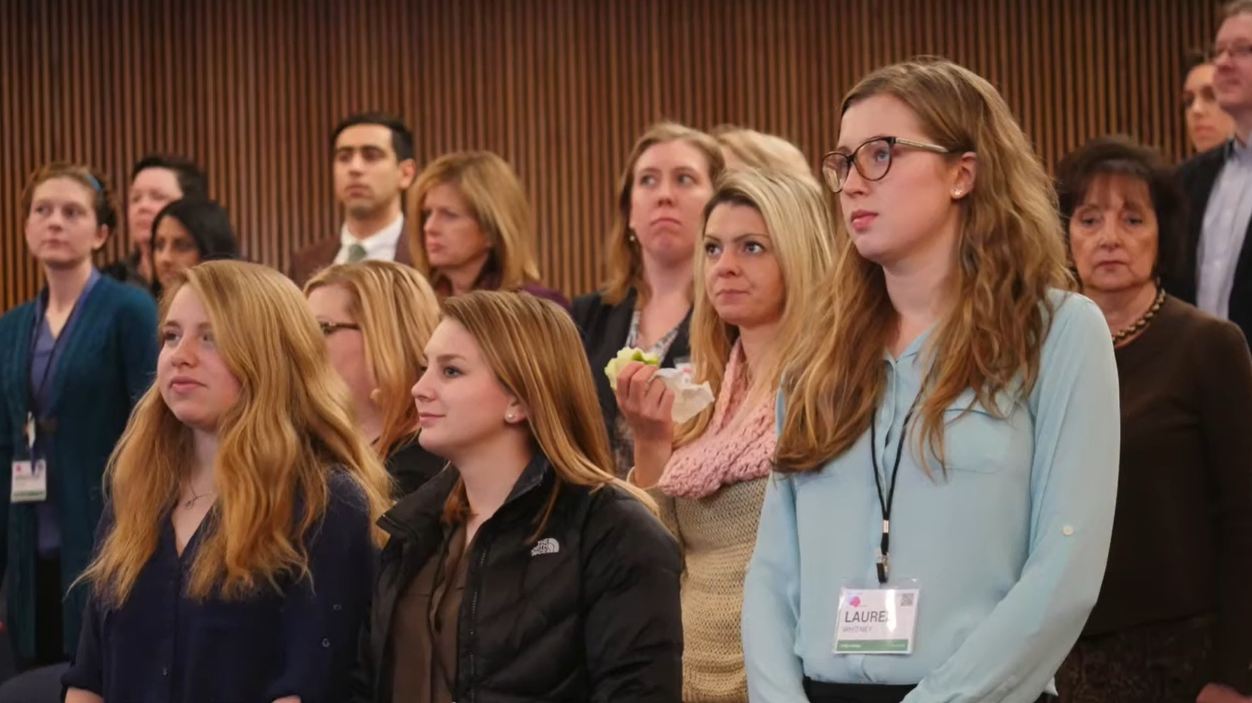 Group of people attending a conference or seminar, standing and listening attentively, with some wearing name badges.