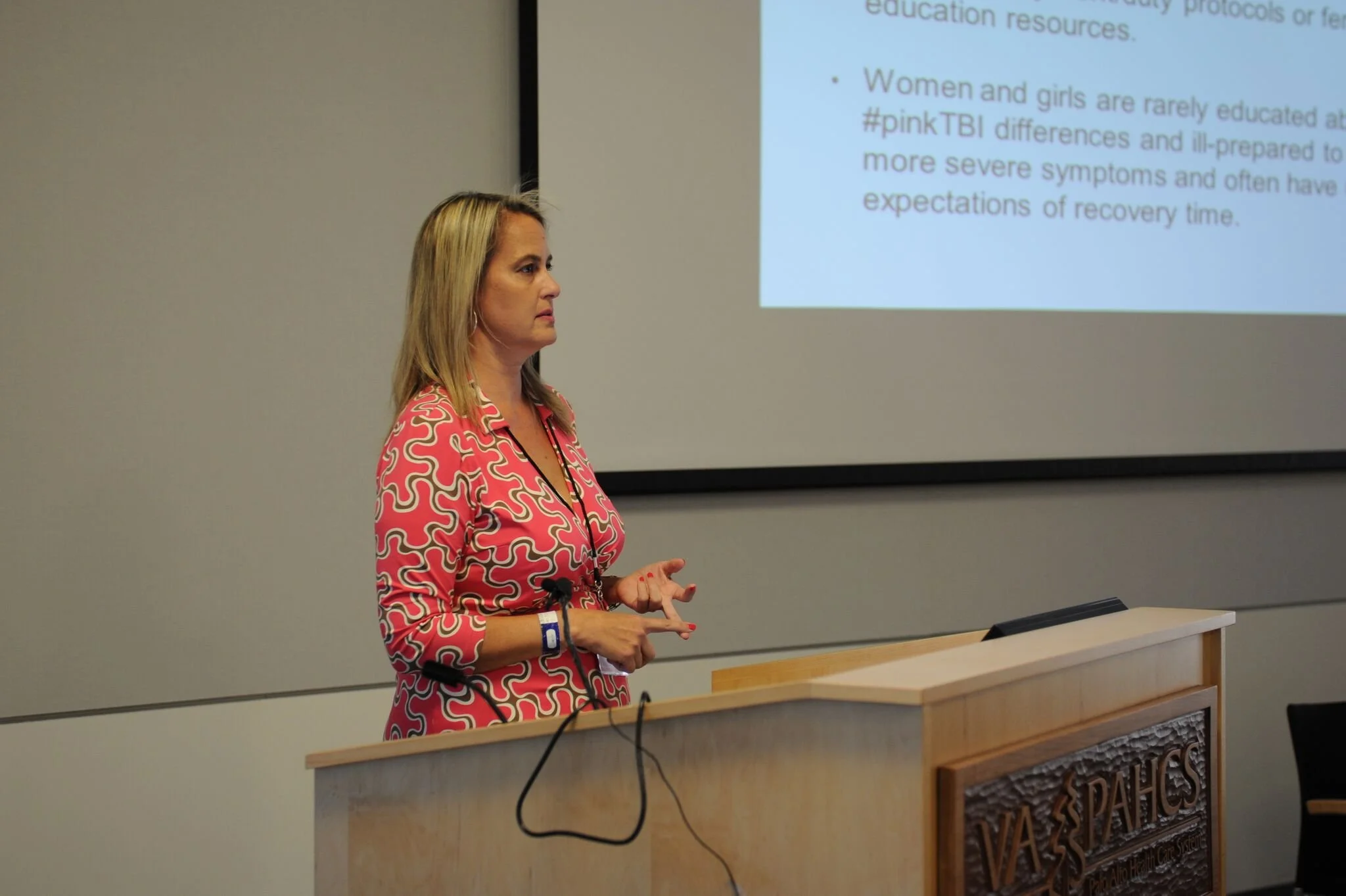 A blonde woman in a pink patterned shirt standing at a podium during a presentation, with a slide projected on the wall behind her showing text about women's education and health issues.