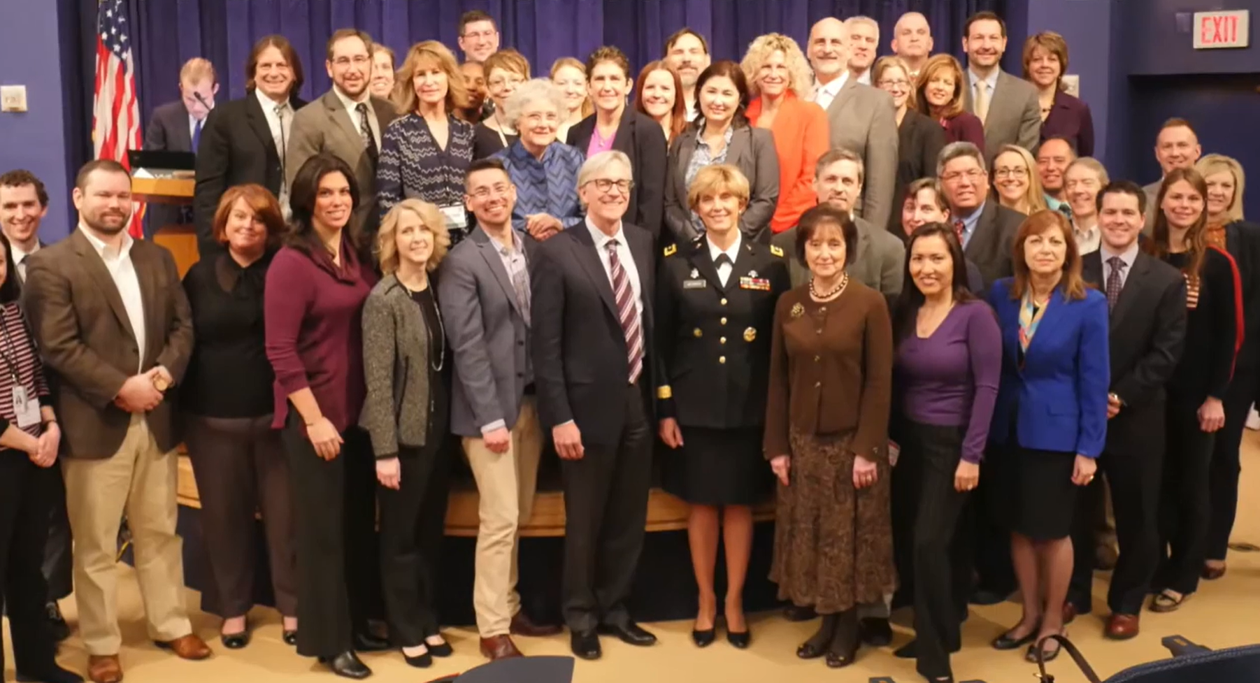 Group of diverse men and women dressed in business attire, standing and smiling for a photo in a conference or meeting room, with a stage, blue curtains, and an American flag in the background.