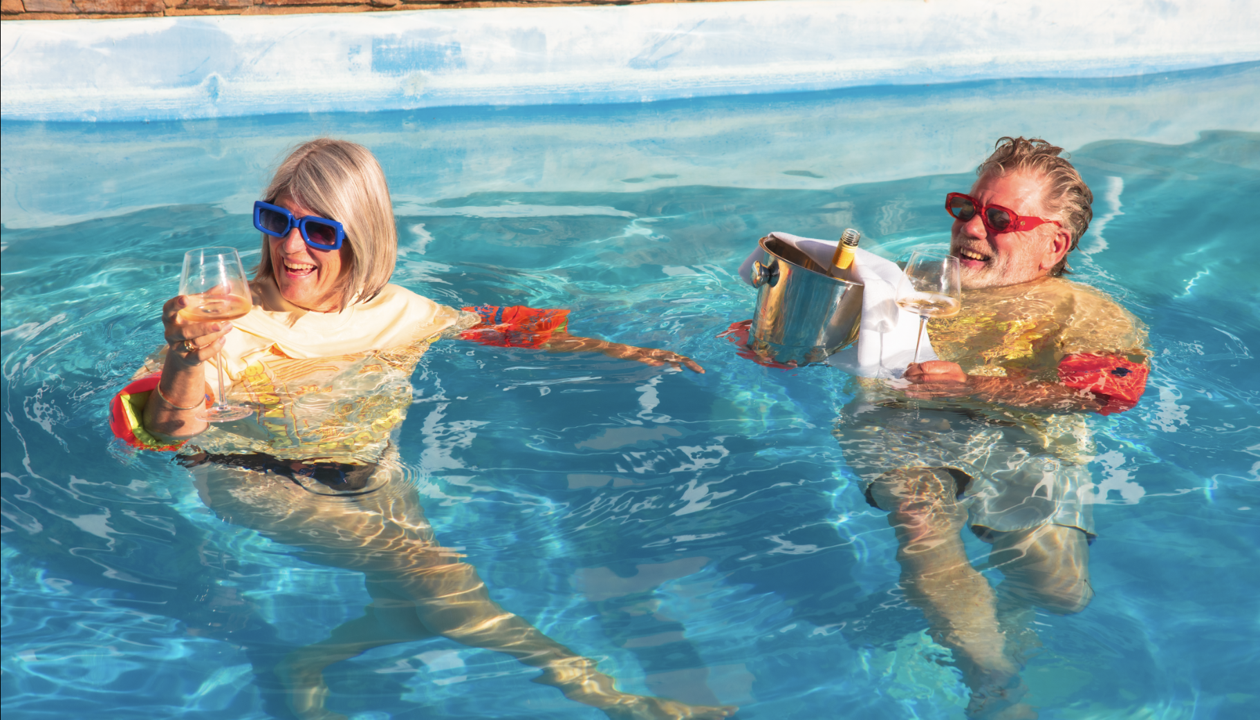 Two seniors in a swimming pool, wearing colorful sunglasses, smiling and holding glasses of wine, with one holding a bucket of ice with a bottle and a glass inside.