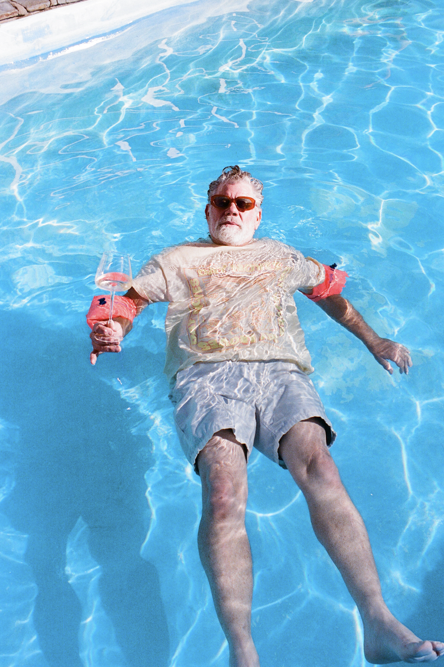 An elderly man with gray hair, beard, and sunglasses floating on his back in a swimming pool, holding a wine glass in one hand.