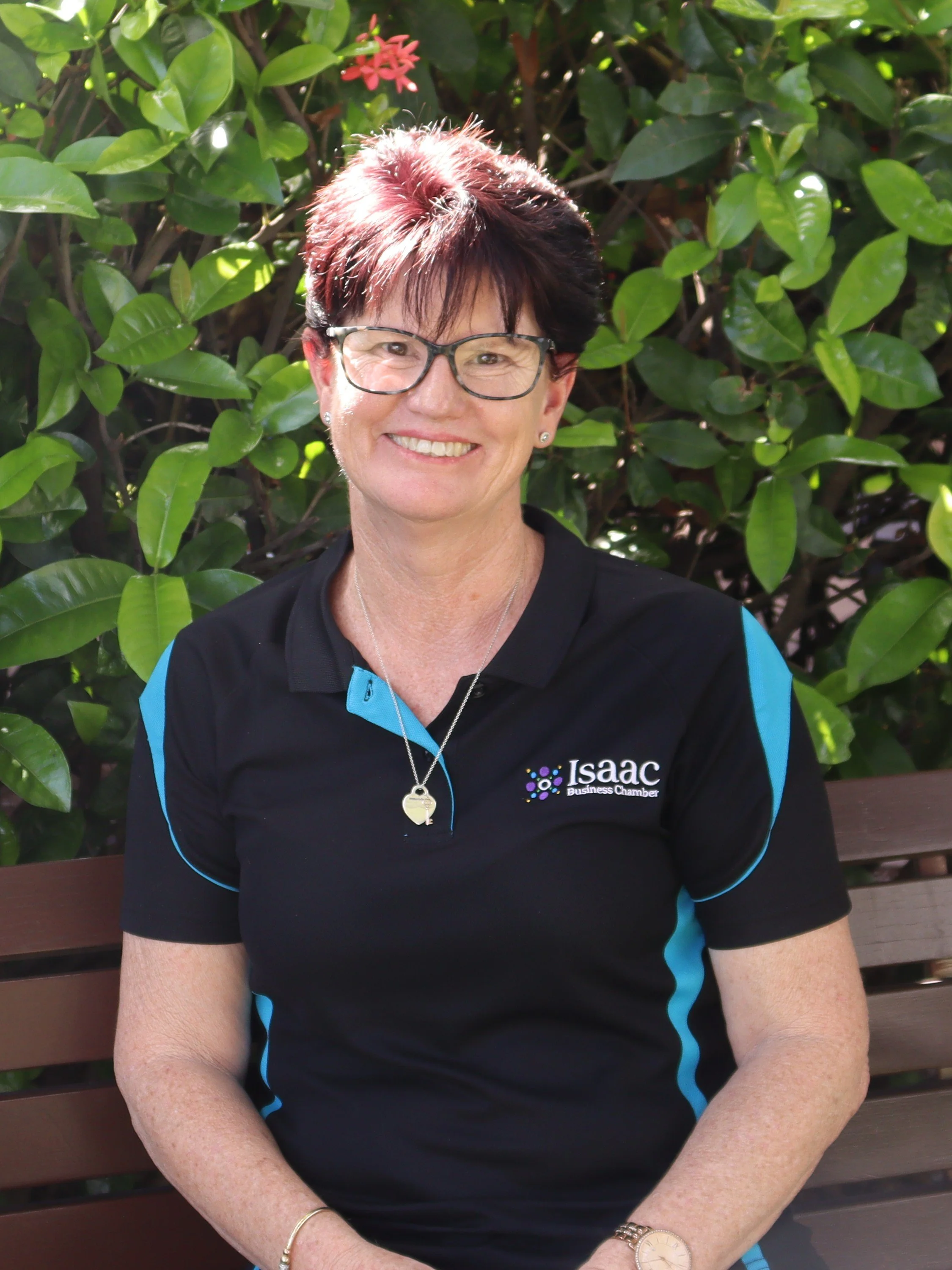A woman with short dark hair, wearing glasses, smiling, dressed in a patterned dress, standing in front of white horizontal slat walls with greenery in the background.