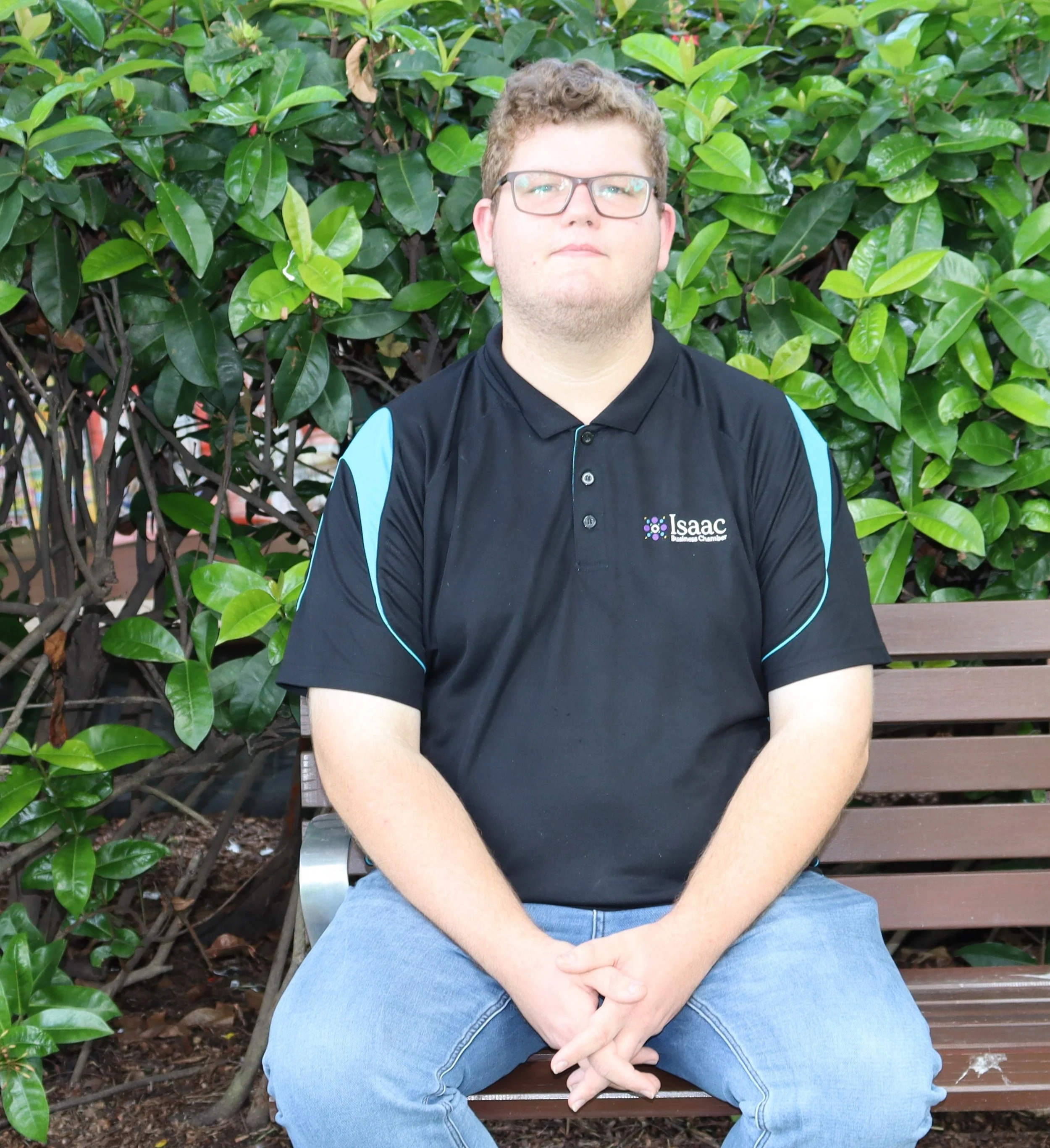 A young man with short brown hair and glasses smiling outdoors with a large tree and green leaves in the background.