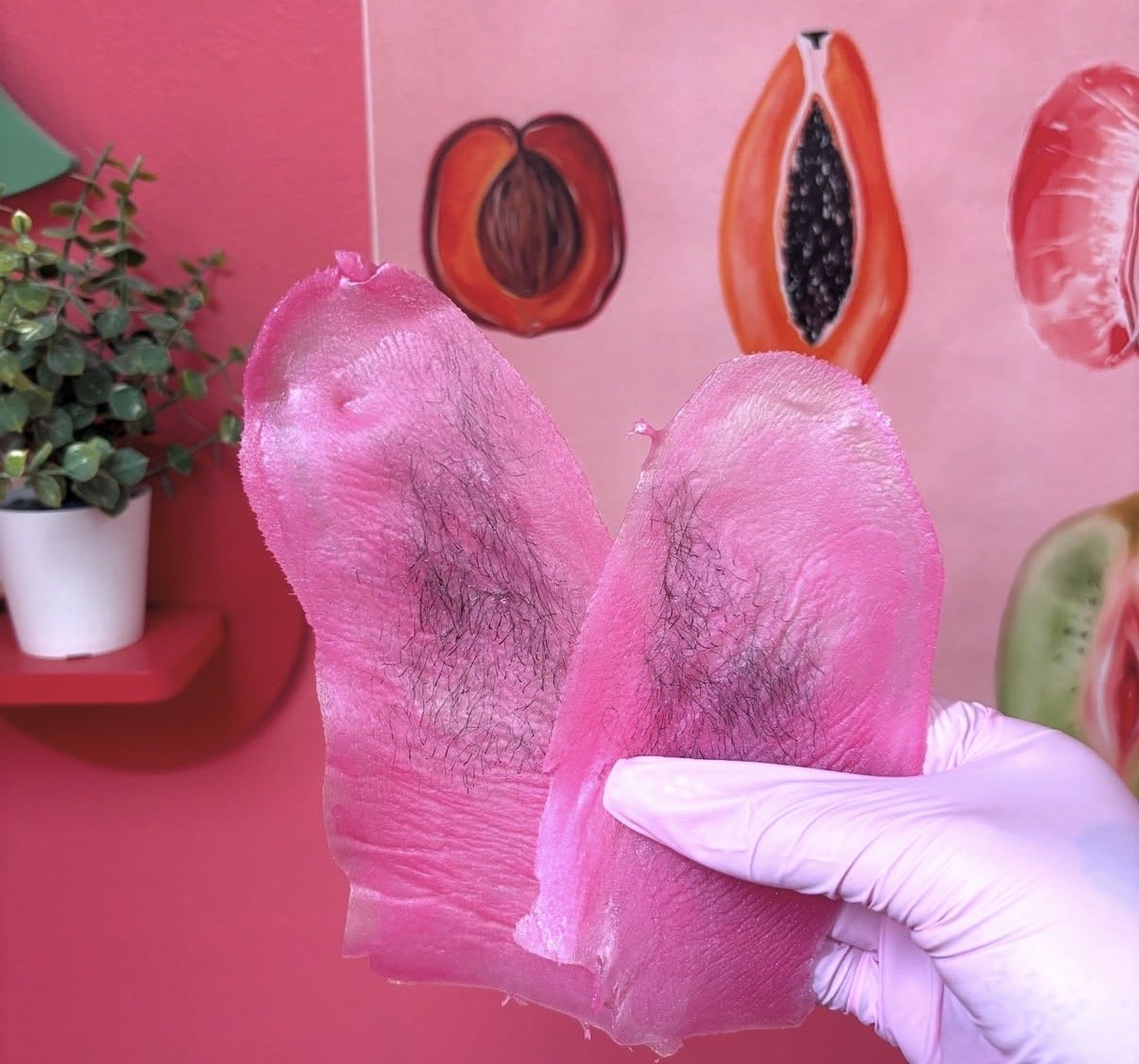 A person with tattoos on their arms is removing a gel nail extension from their finger using a wooden cuticle stick. The person is wearing multiple rings and a green jade bracelet, and has a green background with blurred pink and yellow stripes.