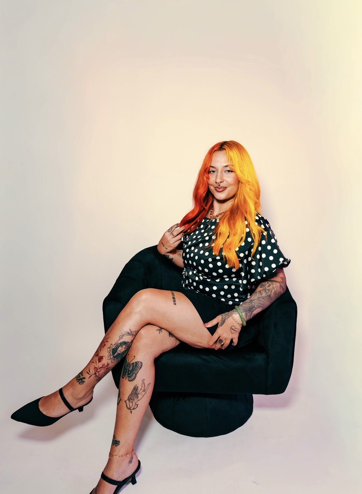 A woman with bright pink and yellow hair sitting on a black leather chair against a light blue background. She is wearing a black pinstripe blazer and has tattoos on her hands. She is smiling subtly, looking directly at the camera.