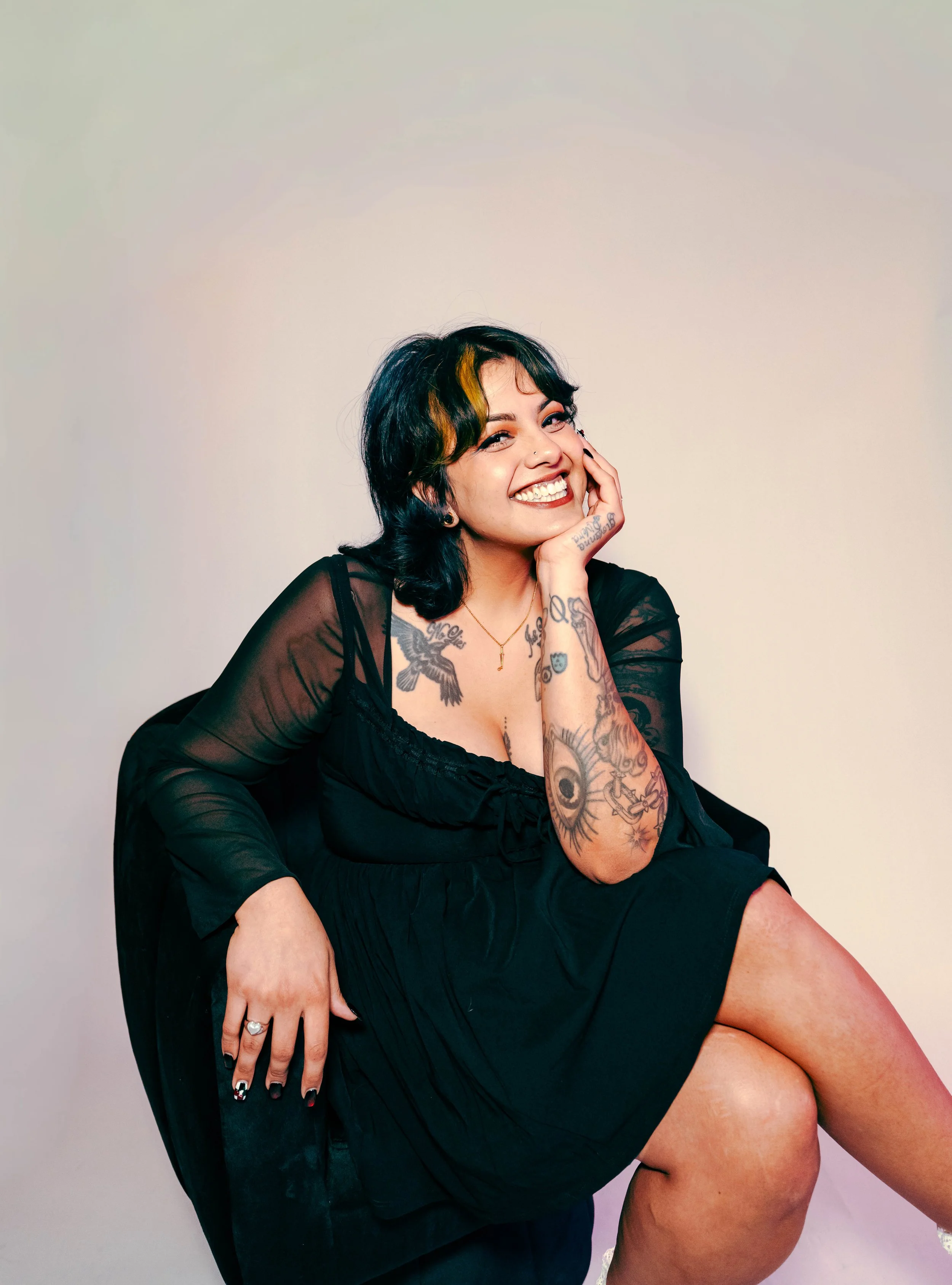 A woman with black and red hair, wearing a colorful, patchwork-style dress with a plaid pattern, sitting on the floor against a plain white background and smiling.