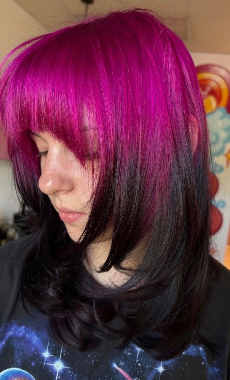 Close-up of a woman with shoulder-length hair dyed in multiple colors including purple, green, and blue, wearing glasses, with part of her face and shoulder visible.