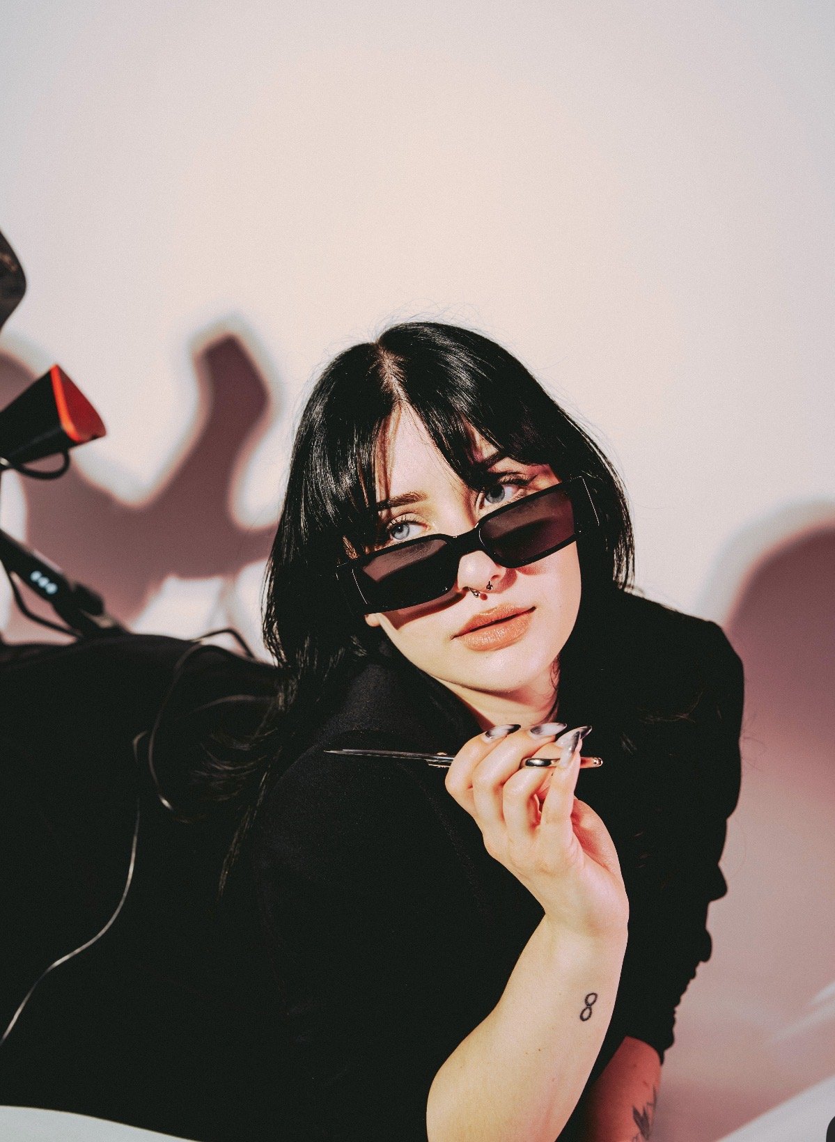 A young woman with black hair, light skin, and tattoos, wearing black sunglasses and a black shirt, posing against a light background.