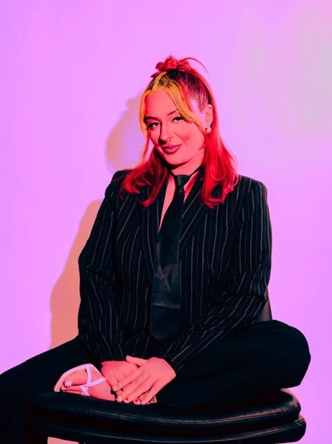 A woman with long pink and yellow hair sitting cross-legged on a black cushion against a pink background. She is wearing a pinstripe black suit with a sheer shirt underneath, smiling at the camera.