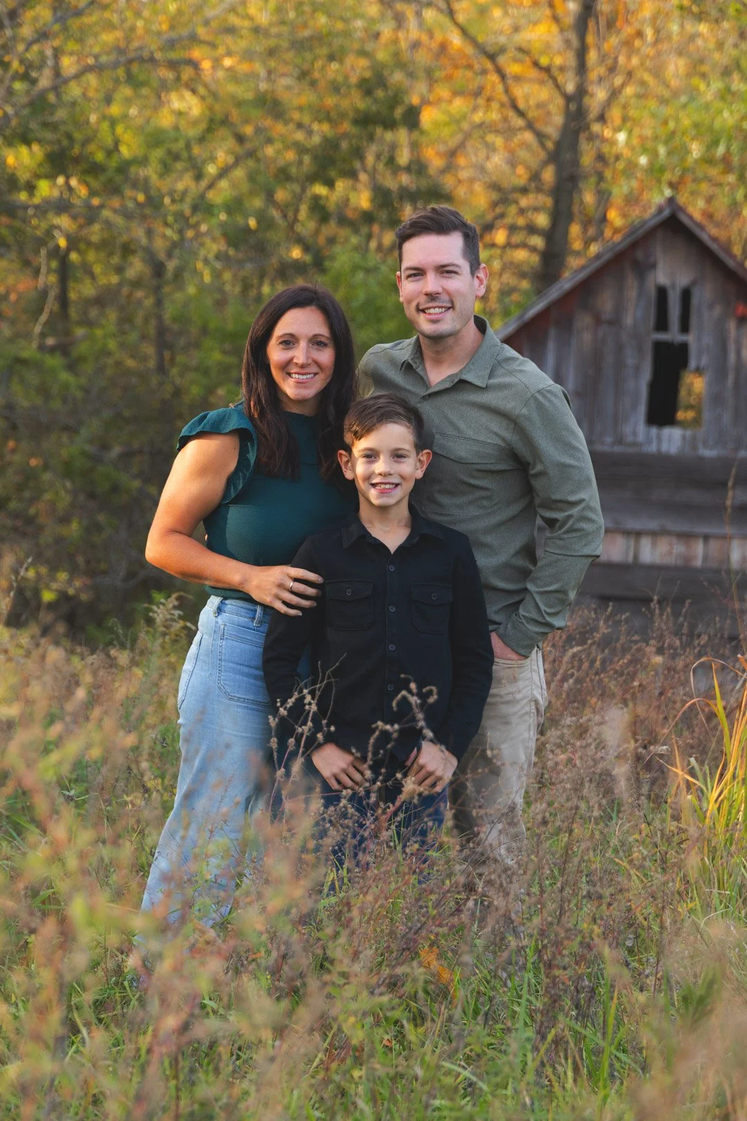 A smiling family of three, including a woman, man, and young boy, standing outdoors in a field with tall grass and wildflowers, with autumn-colored trees and a rustic wooden barn in the background.