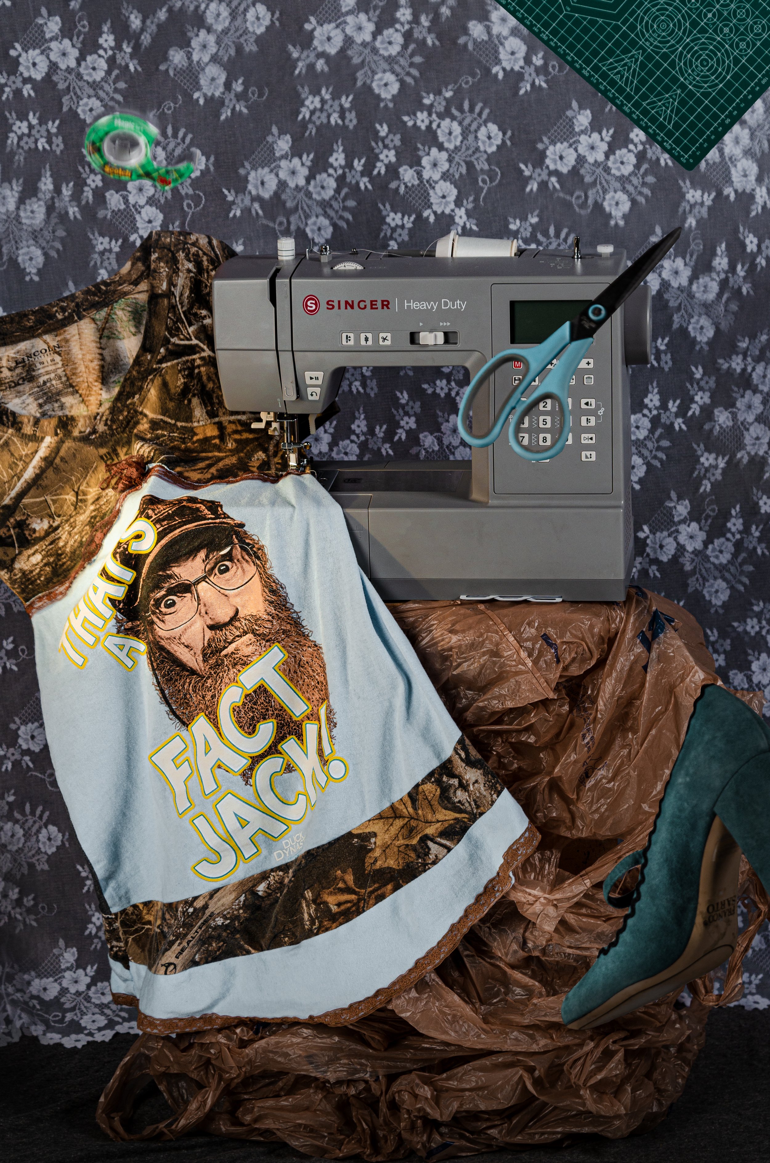 A sewing machine on a table covered with brown plastic and fabric, with a humorous T-shirt featuring a man's face and the words 'FAC JACKET!', a pair of scissors hanging over the machine, a sewing tape measure, and a cutting mat on top, all against a