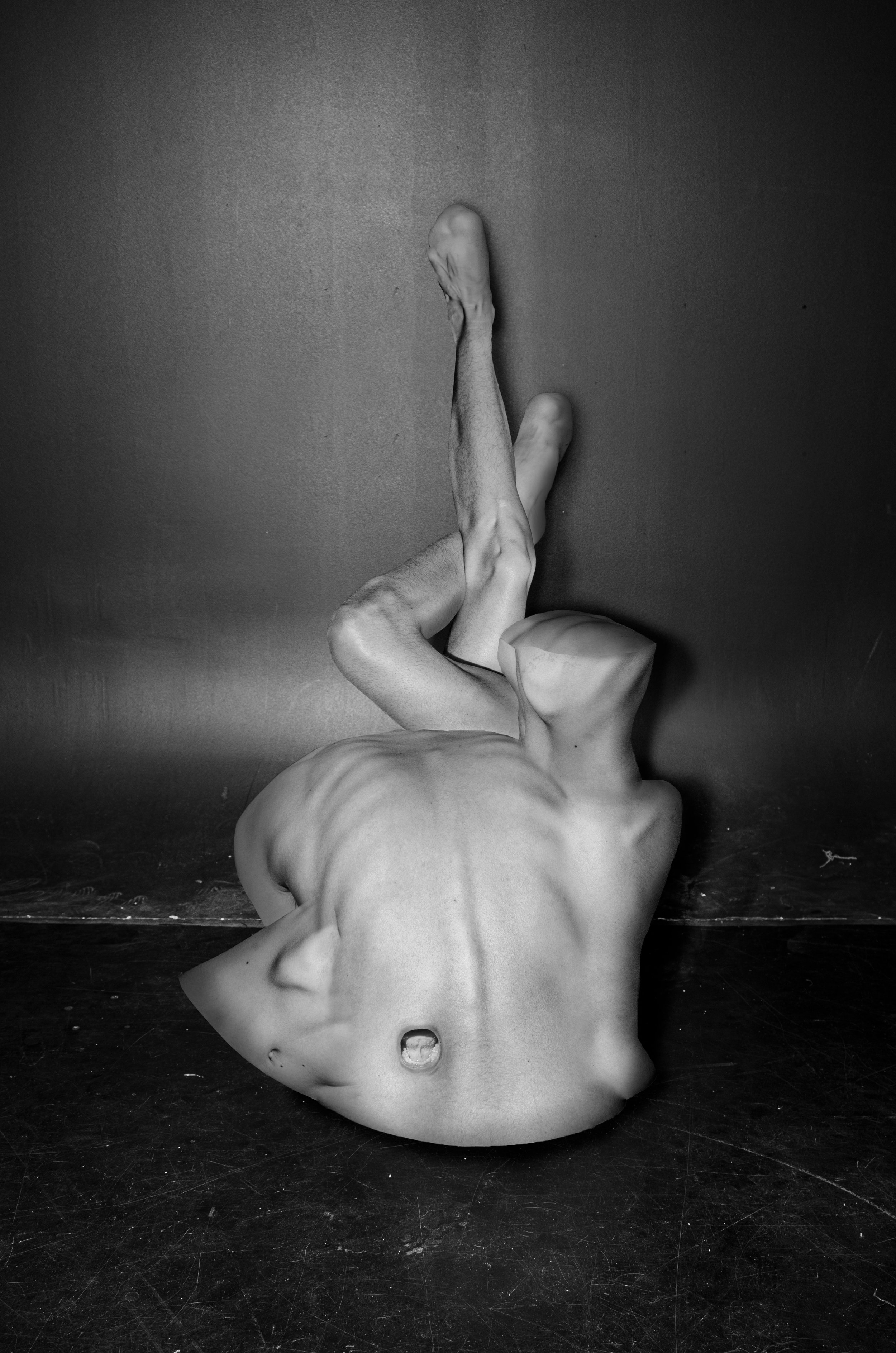 A black and white sculpture of a human torso, headless and with no arms, positioned on the ground. The sculpture's body is upside down, with legs extended upward and crossed at the ankles.