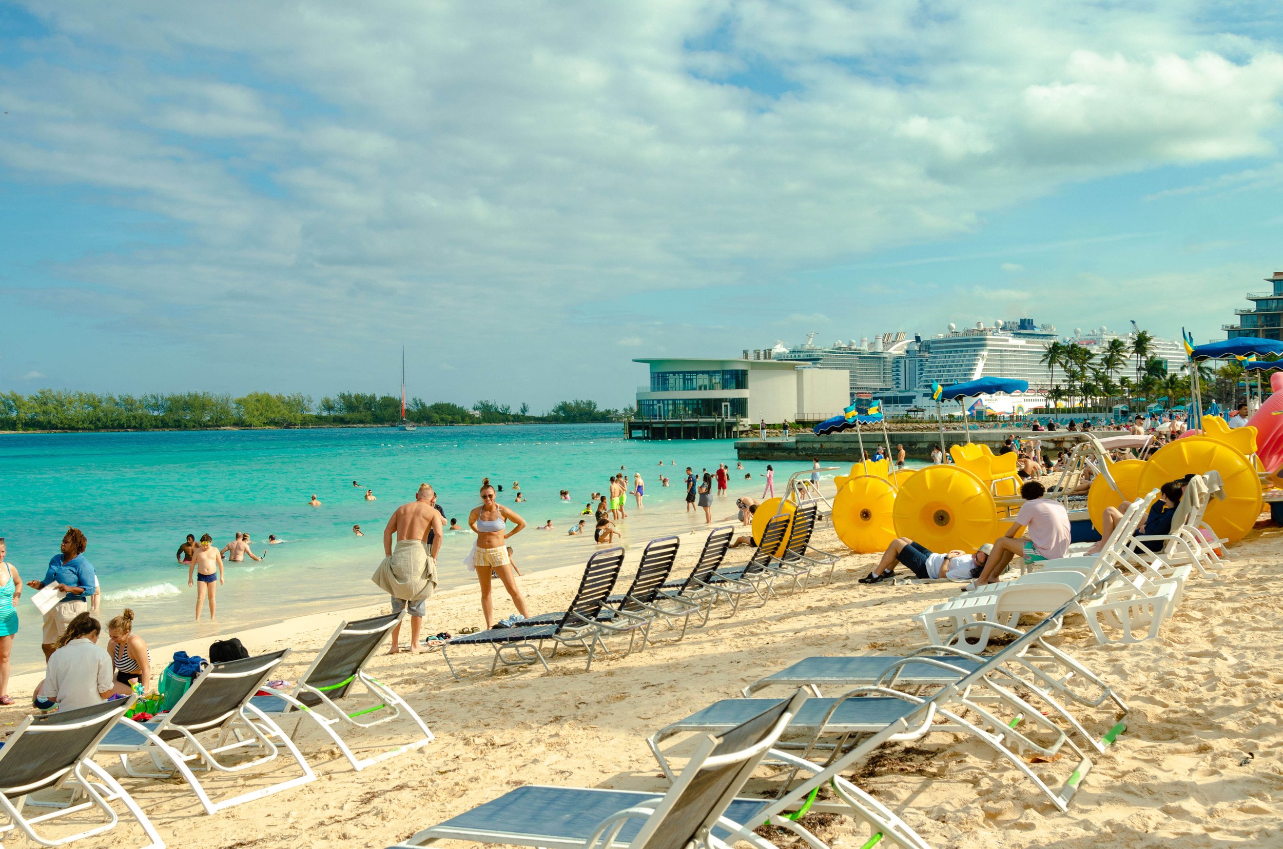 Beach scene with lounge chairs, people swimming in clear blue water, and cruise ships docked near the shore, under a partly cloudy sky.