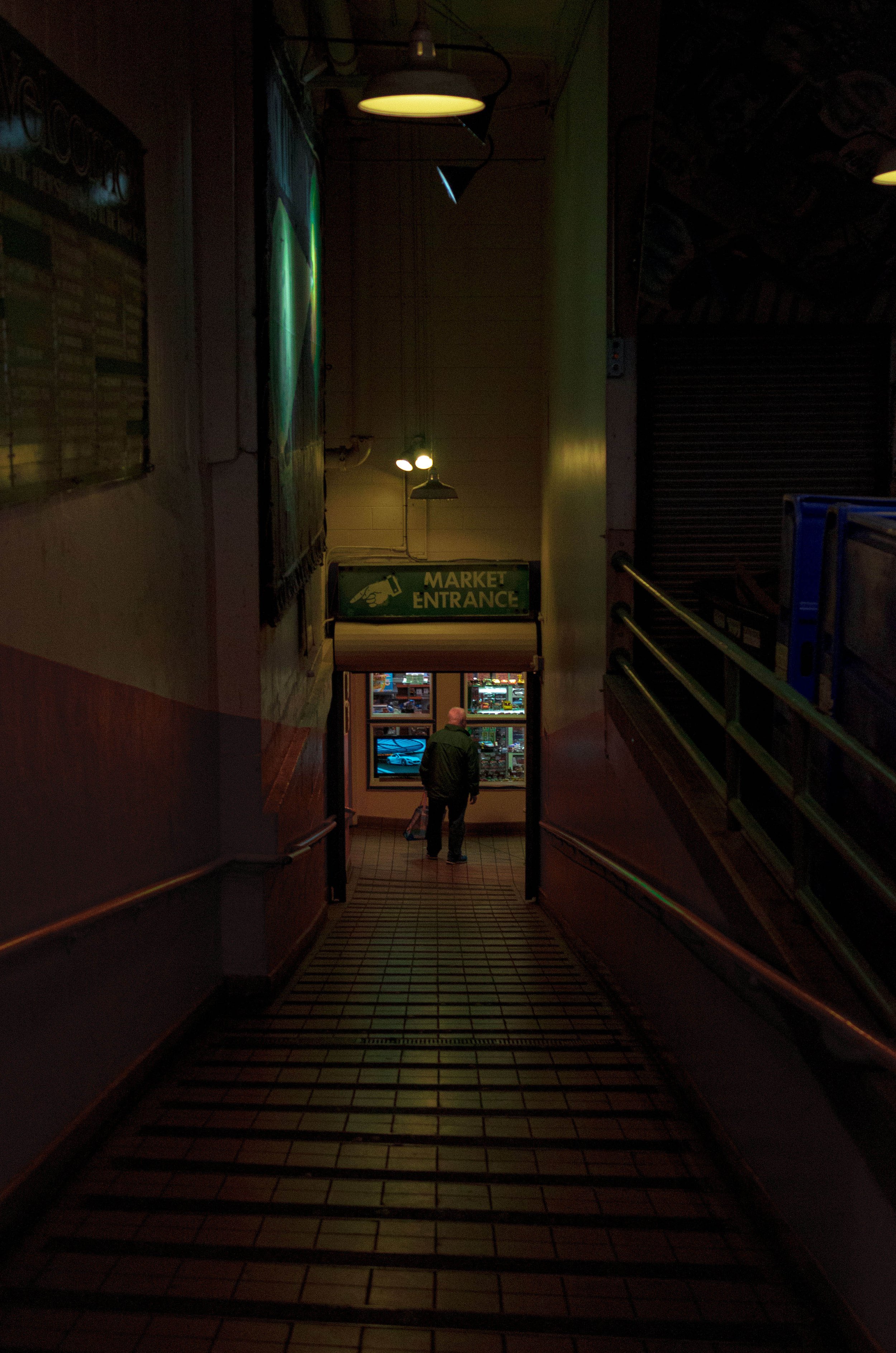 A person standing at the entrance of a market, looking inside. The sign above says 'Market Entrance'. The scene is dimly lit with a few lights overhead.