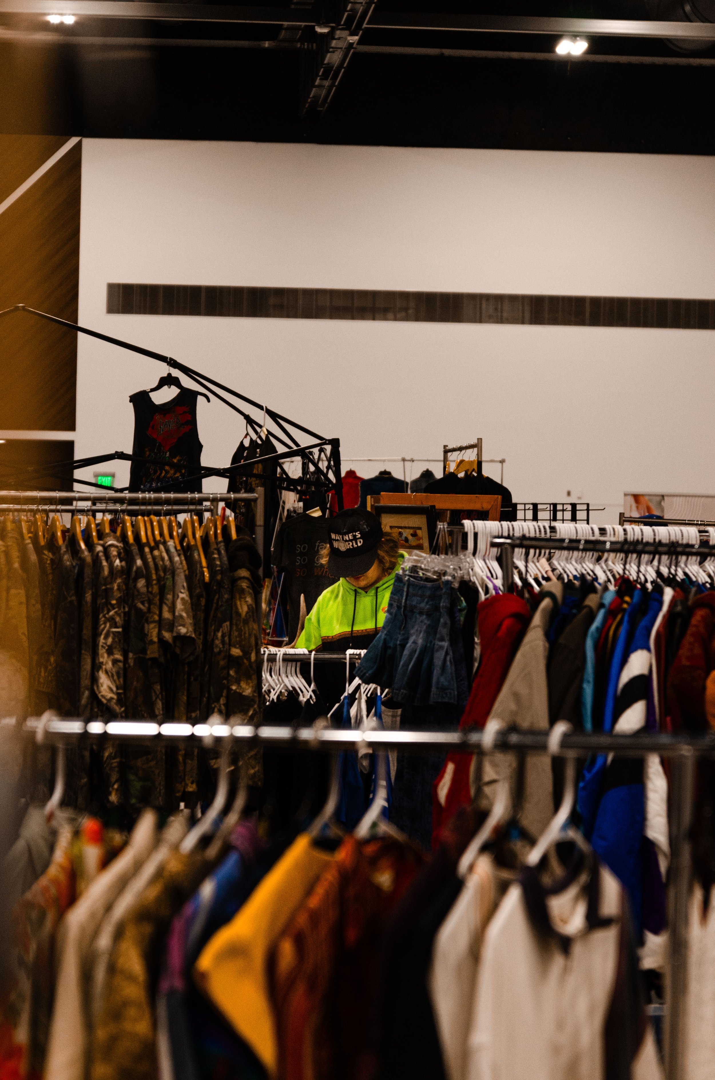 Person browsing racks of clothing in a thrift store or warehouse, wearing a bright yellow-green jacket and a black hat.