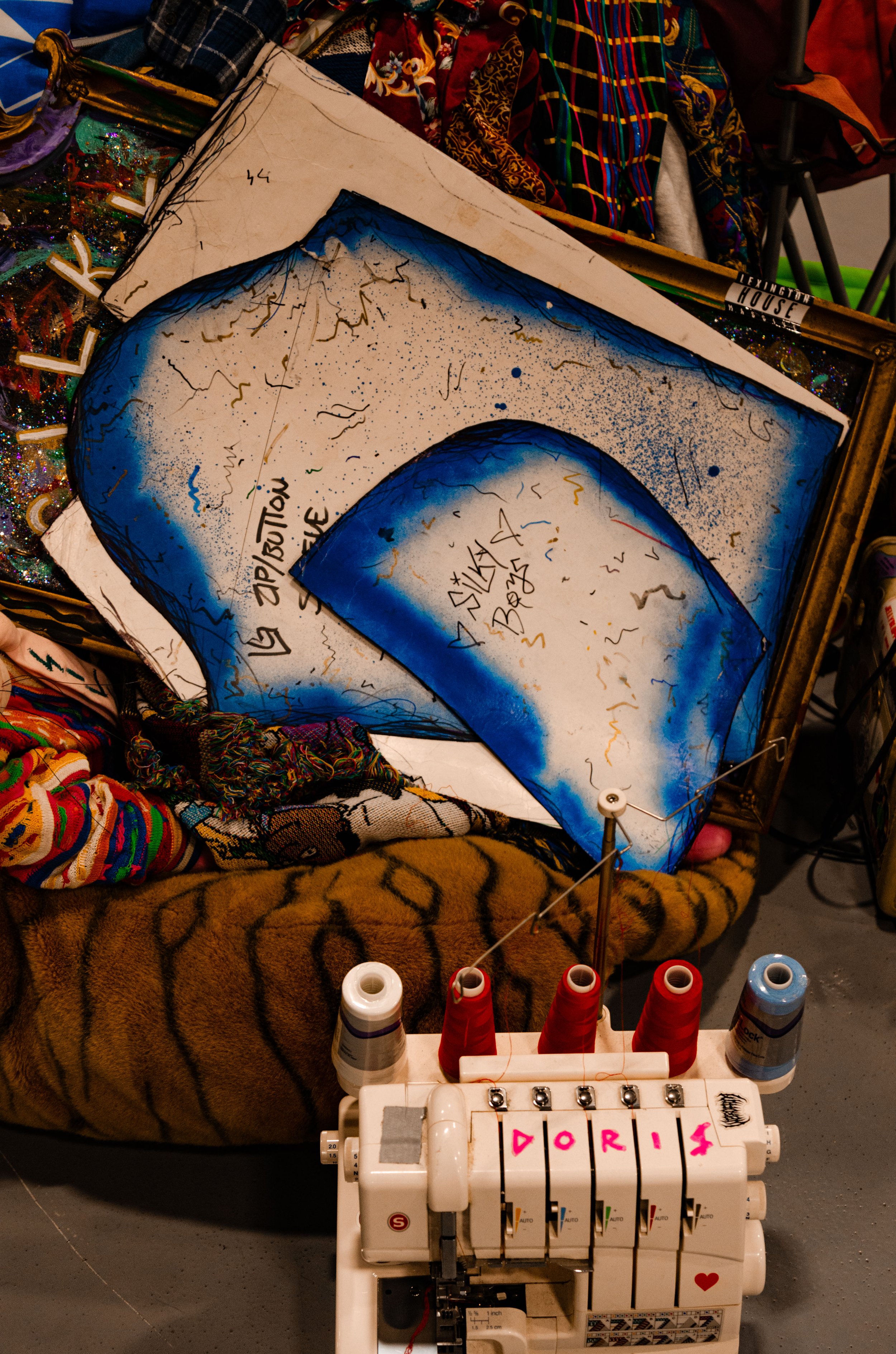 Colorful handmade or painted fabric art pieces with blue and white color scheme, some featuring text and abstract design, displayed among sewing supplies including spools of thread with 'Doris' written on them, and fabric in patterns like tiger print