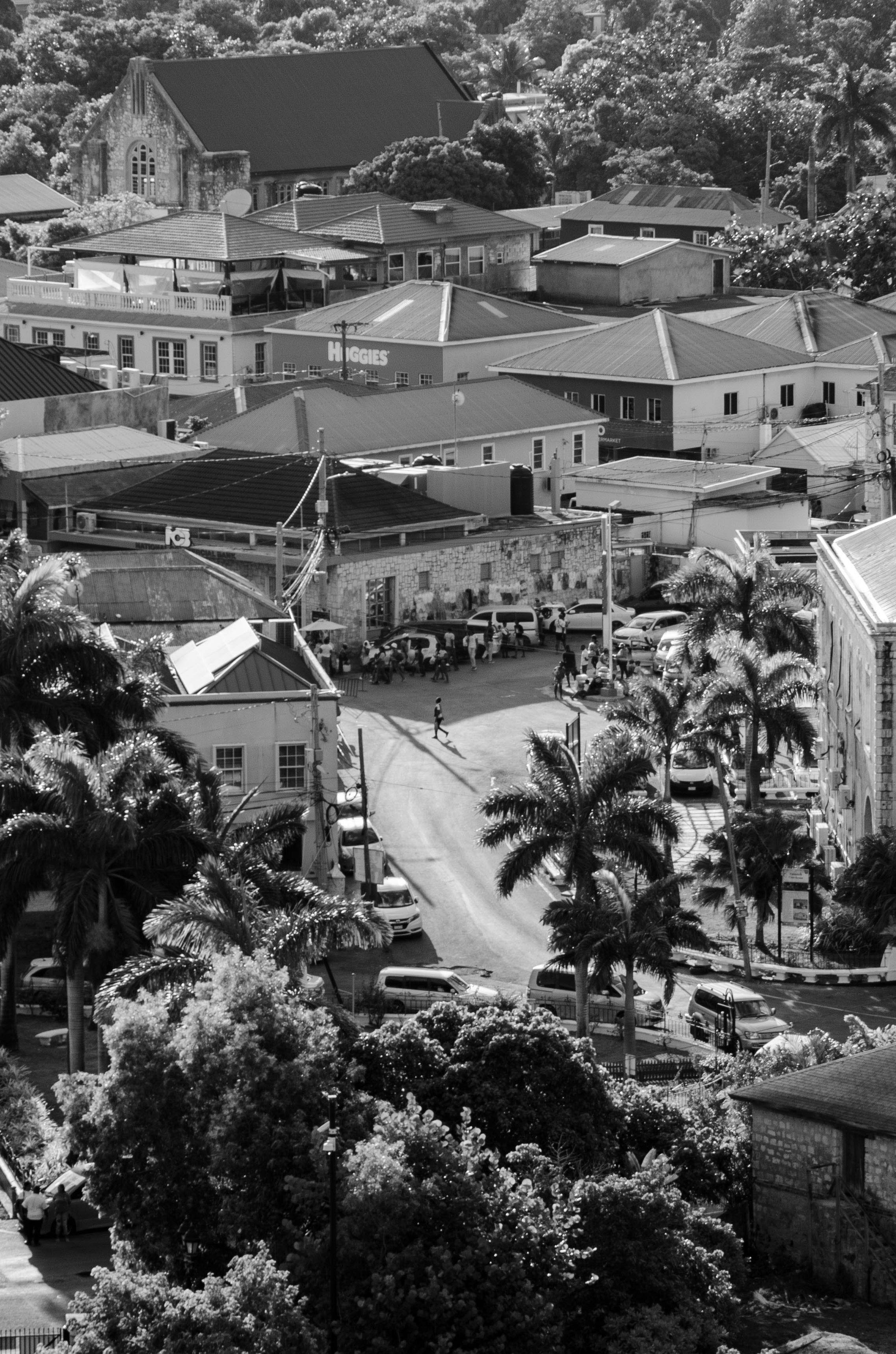 Black and white photo of a hillside neighborhood with tightly packed buildings, palm trees, and a street with parked cars, people walking, and a small gathering near a building.