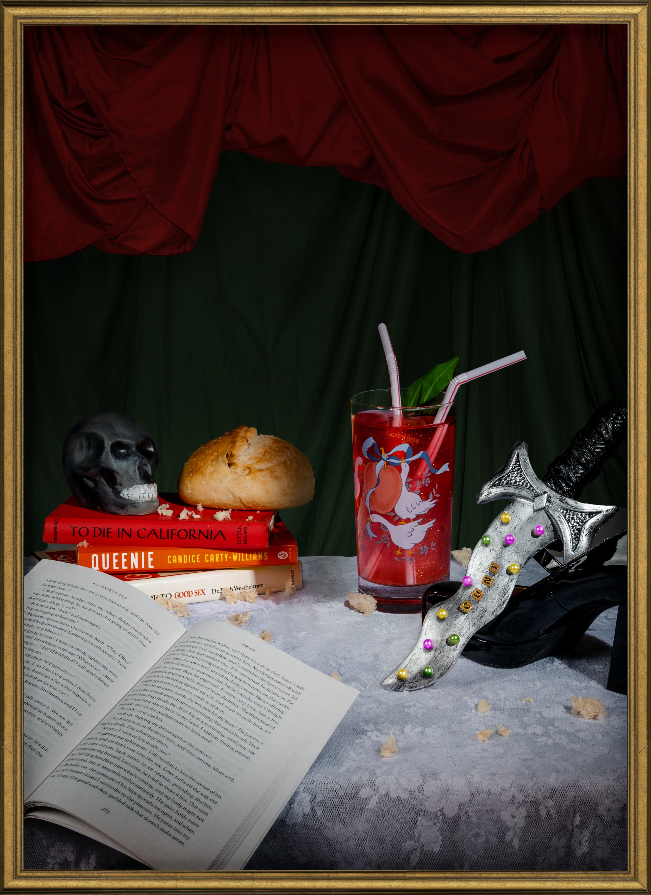An artistic still life featuring a red curtain backdrop, a table with a lace tablecloth, an open book, a stack of books including "To Die in California" and "Queenie," a black skull, a bread roll, a red drink with straws and fruit, a decorative dagge