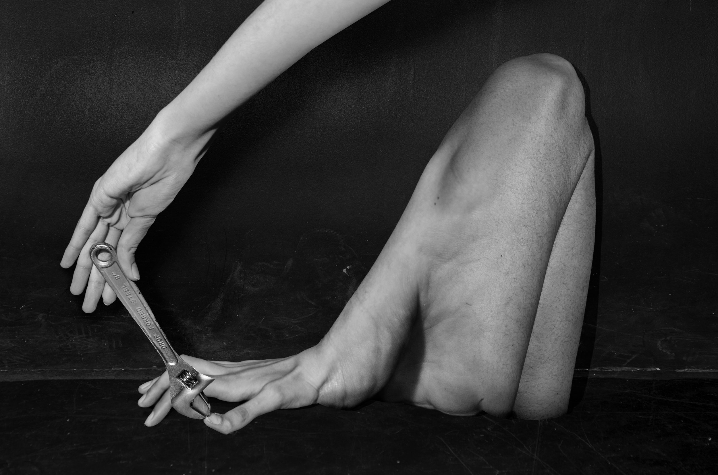 A human arm and leg are positioned to form a loop, with the leg bent at the knee and the arm reaching out to hold a wrench, creating an abstract visual composition in black and white.