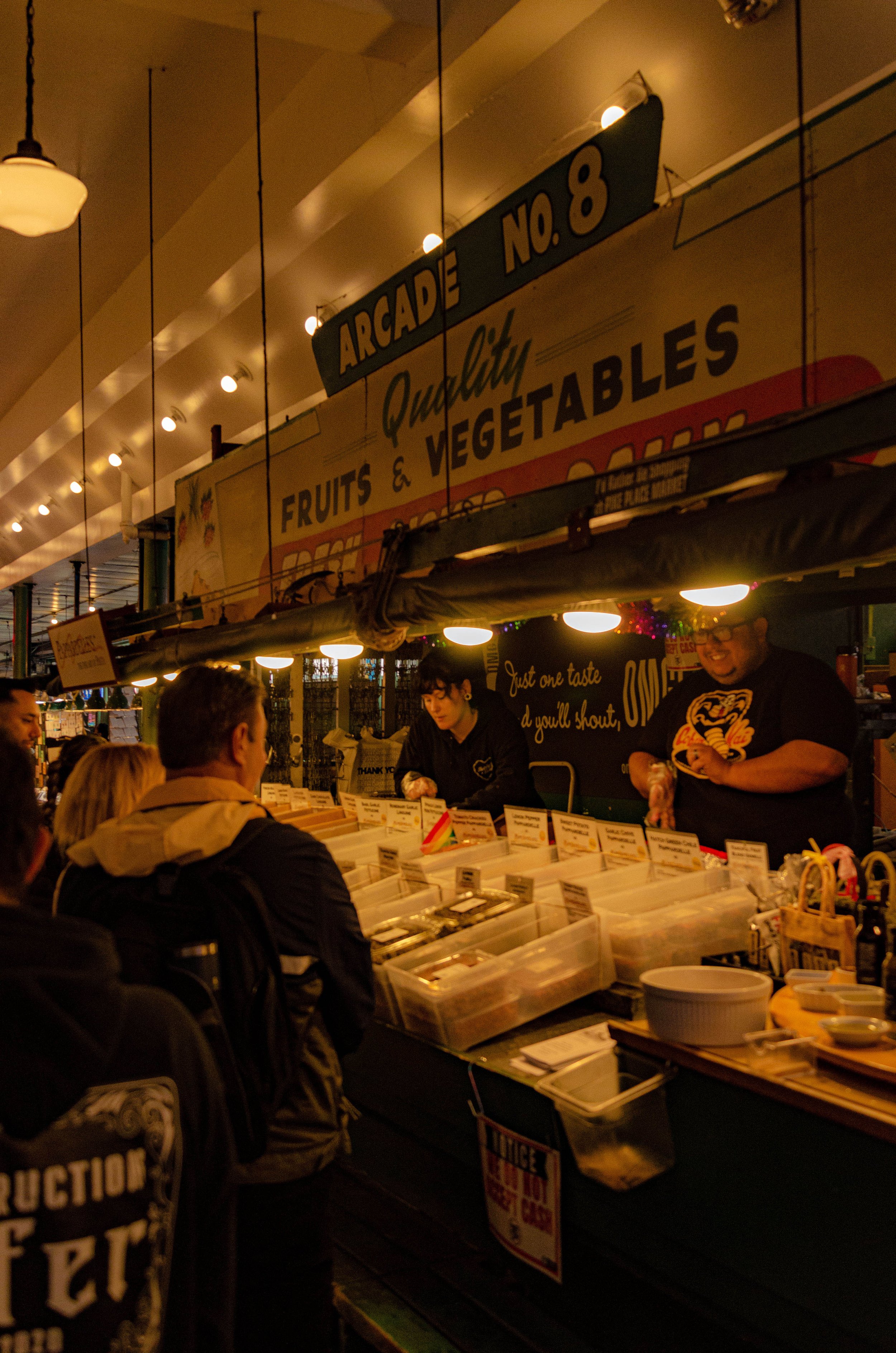 Night scene at a fruit and vegetable market stall with customers shopping and vendors behind the counter, illuminated by warm lights and signs.