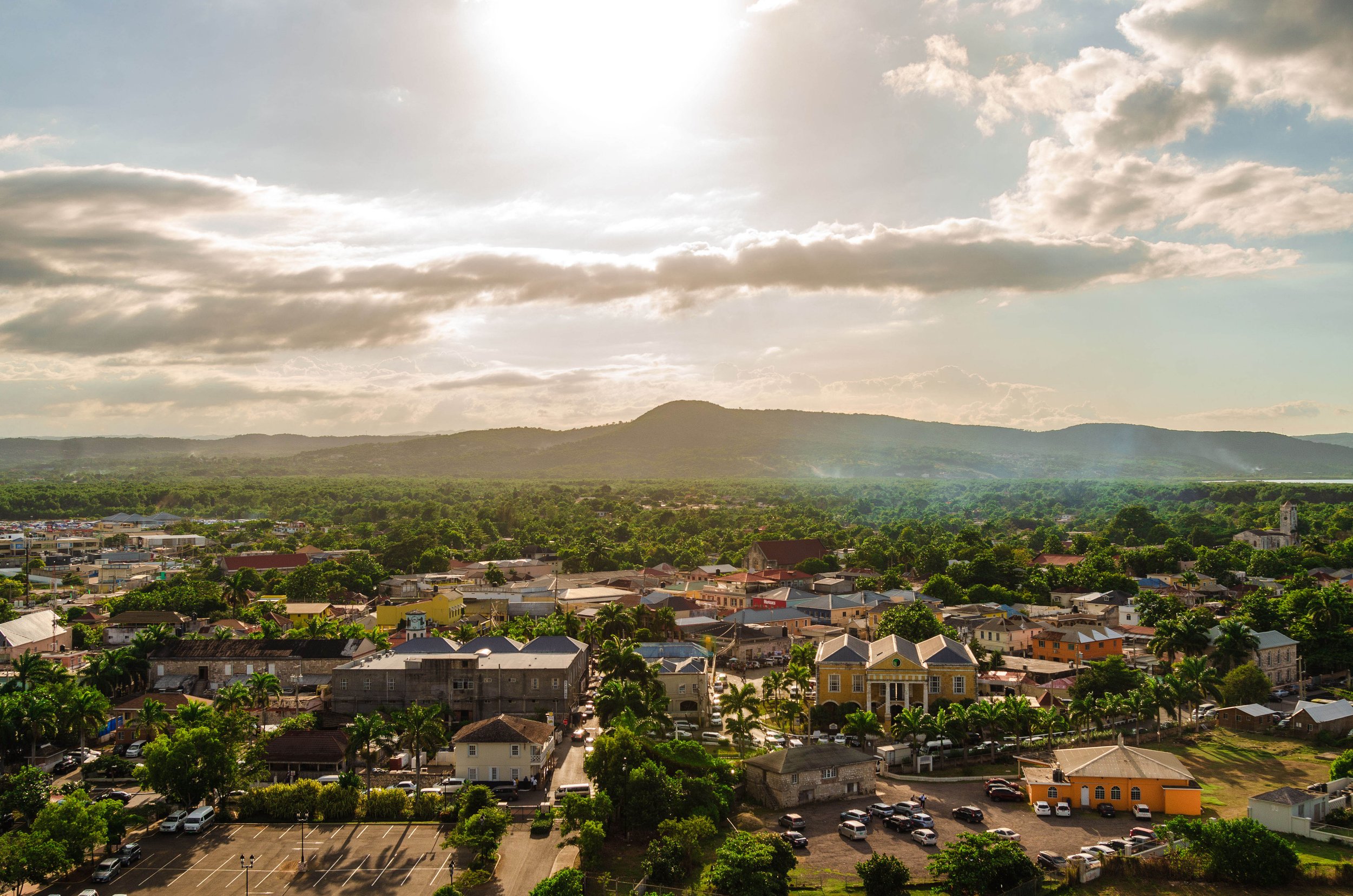 Aerial view of a small town with colorful buildings, surrounded by green trees and a mountain in the background, under a partly cloudy sky.