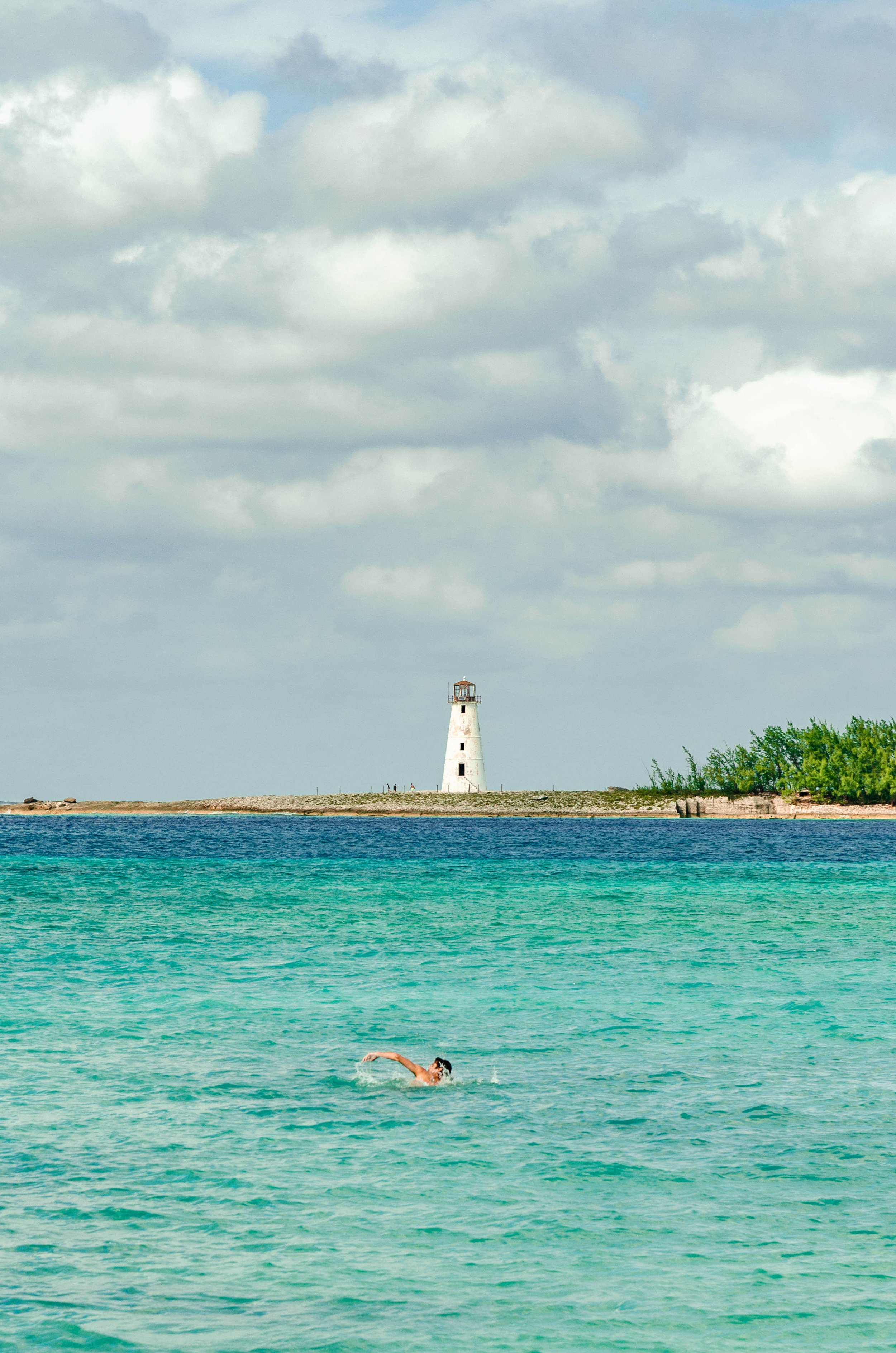 A person swimming in turquoise waters with a lighthouse on the distant shoreline under a cloudy sky.