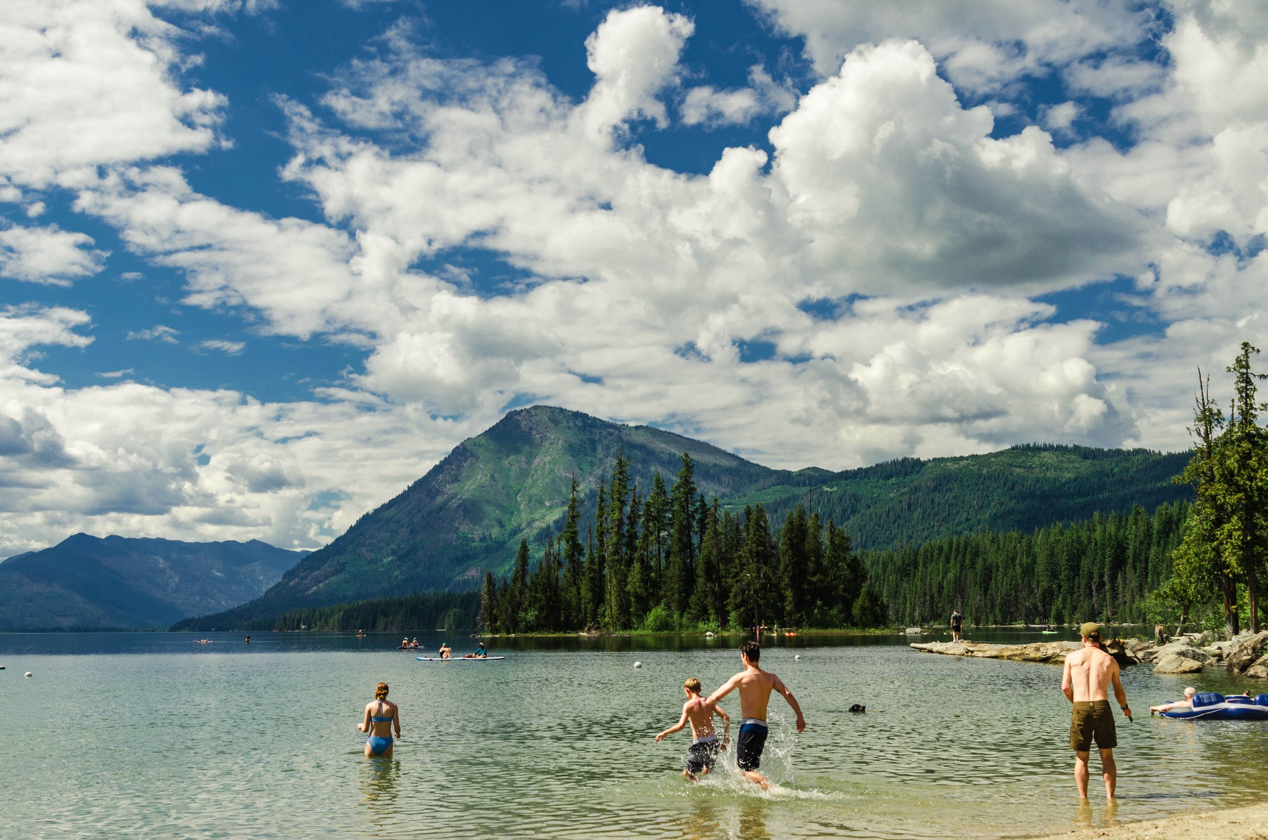 People enjoying a lake with mountain and forest backdrop on a partly cloudy day.