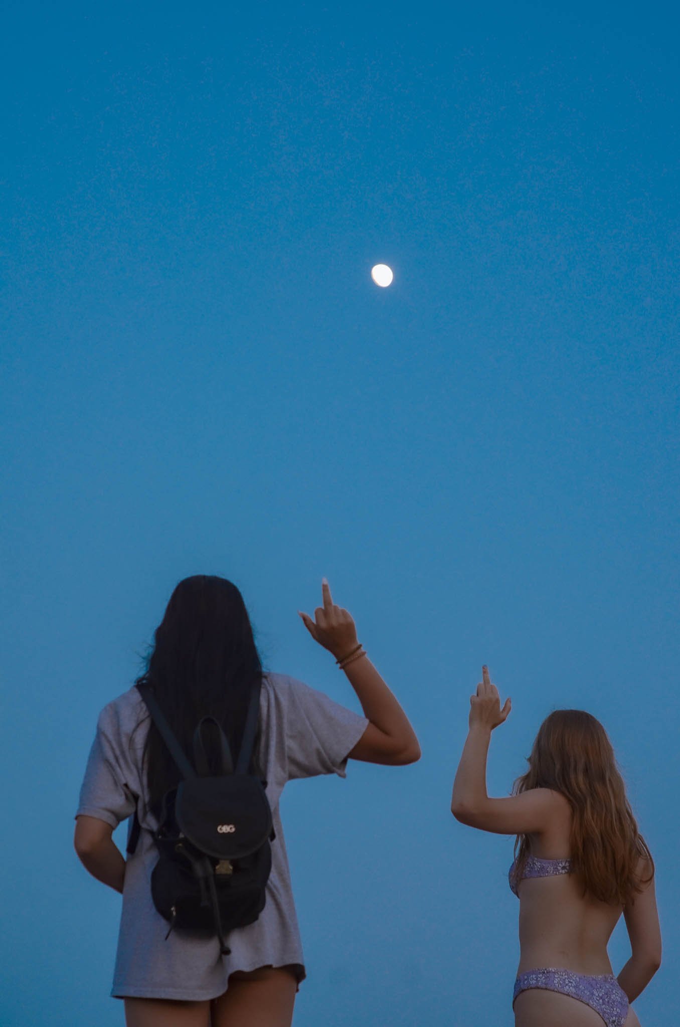 Two women standing outdoors at dusk, pointing at the moon in the sky. One woman wears a grey shirt and backpack, the other wears a bikini top. The sky is clear and blue.