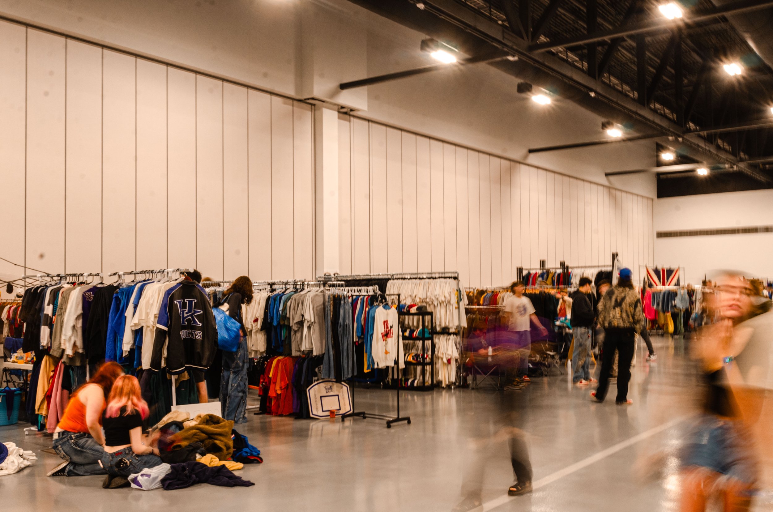 Indoor thrift store with racks of clothes, some people browsing and sorting through items on the floor, and blurred motion of shoppers walking around.