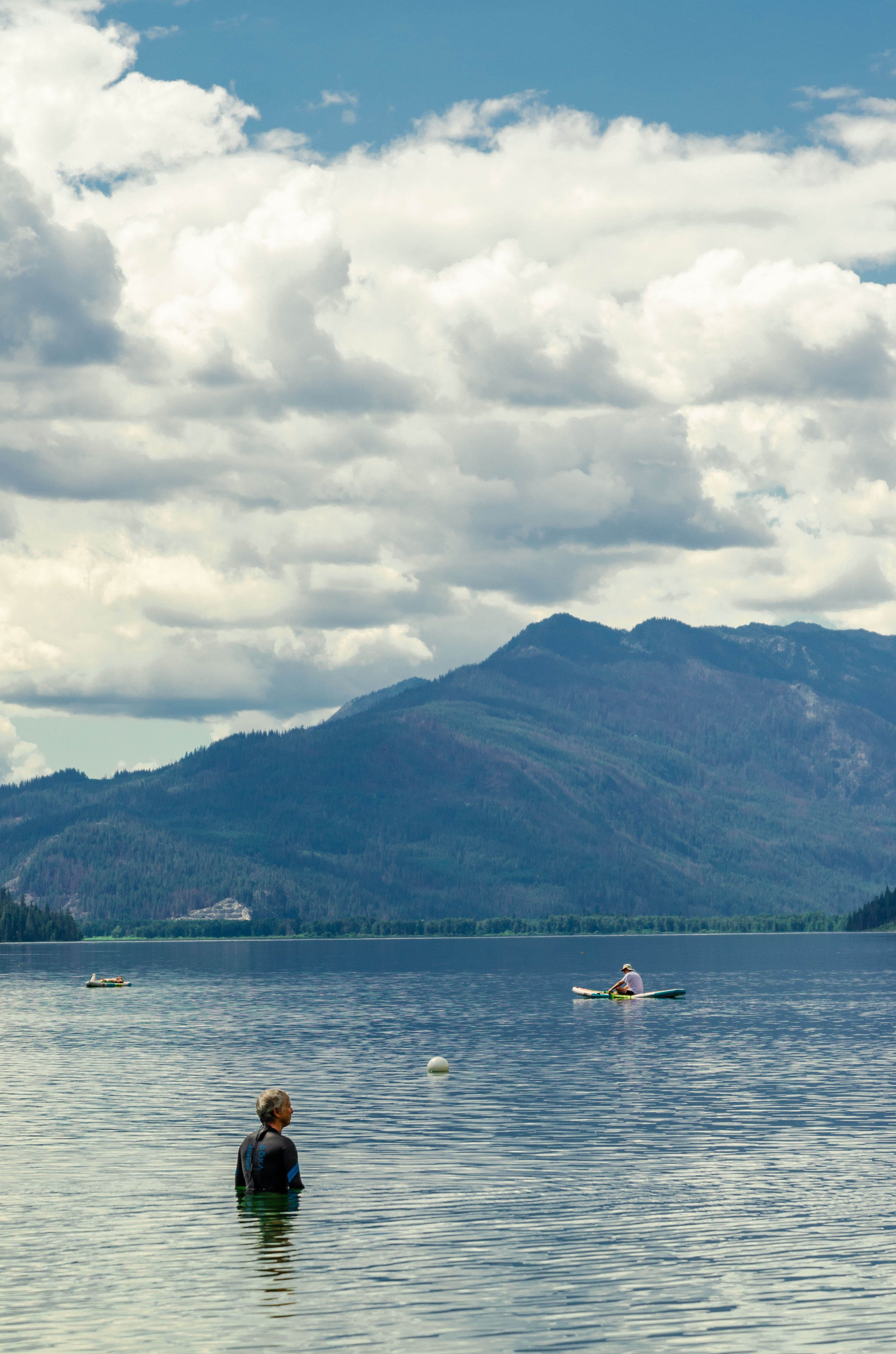 A man stands in a lake with mountains and cloudy sky in the background, while two people kayak in the distance.