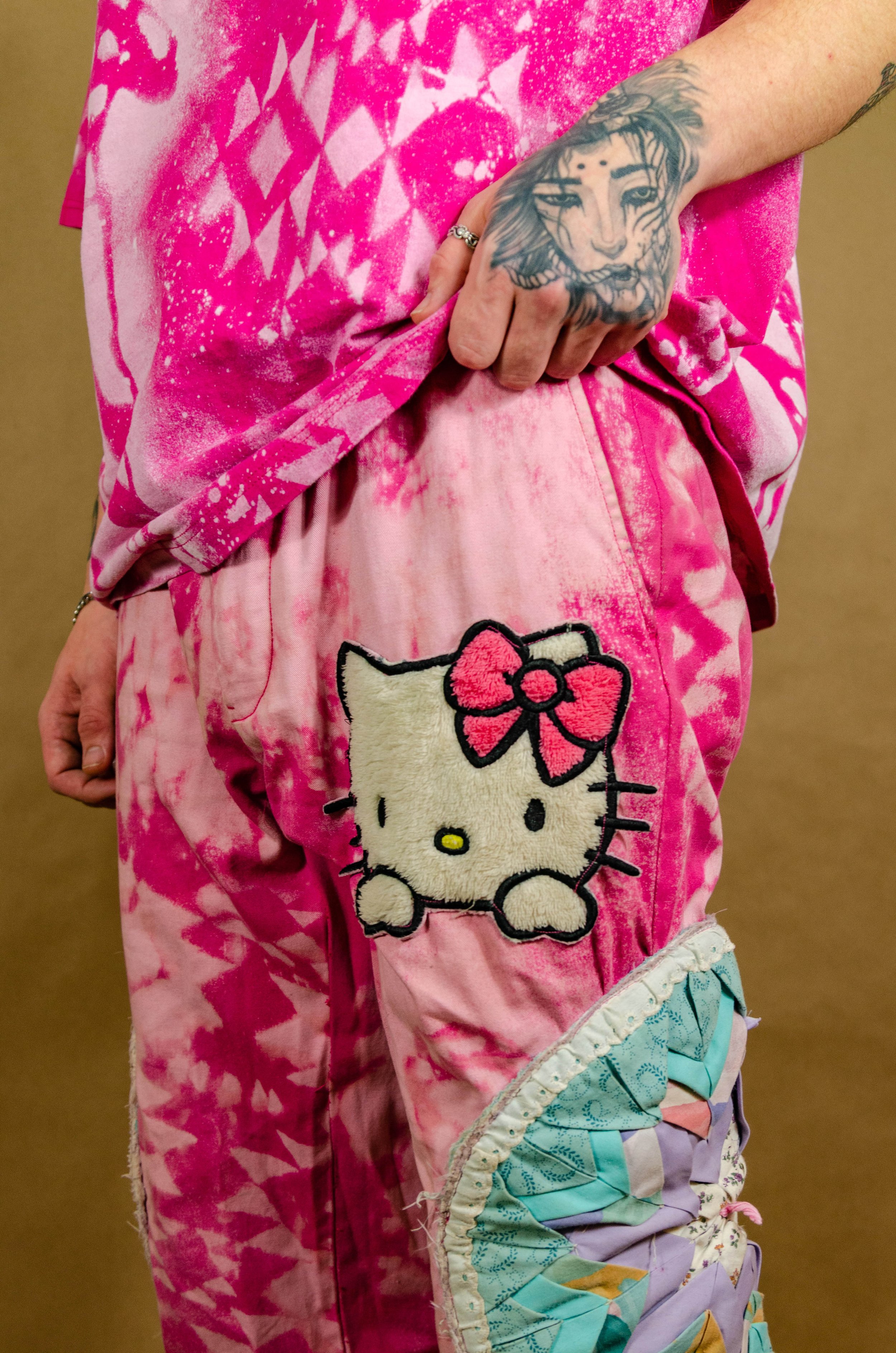 Person wearing pink tie-dye clothing with a Hello Kitty patch on their pants and holding their waist, showing a tattoo of a lion's face on their hand.