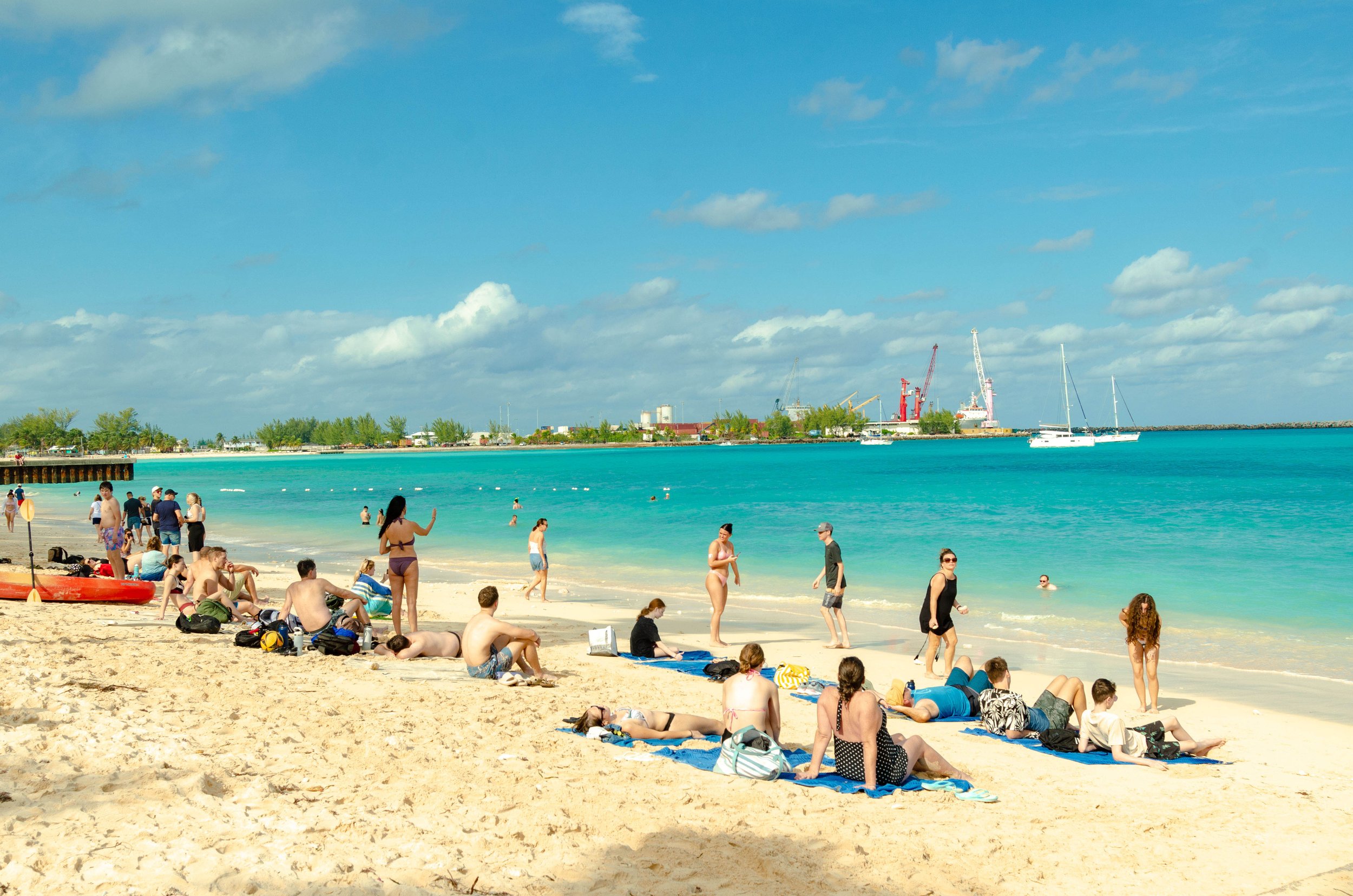 People sunbathing and swimming at a sandy beach with turquoise water, blue sky, and distant industrial cranes and boats