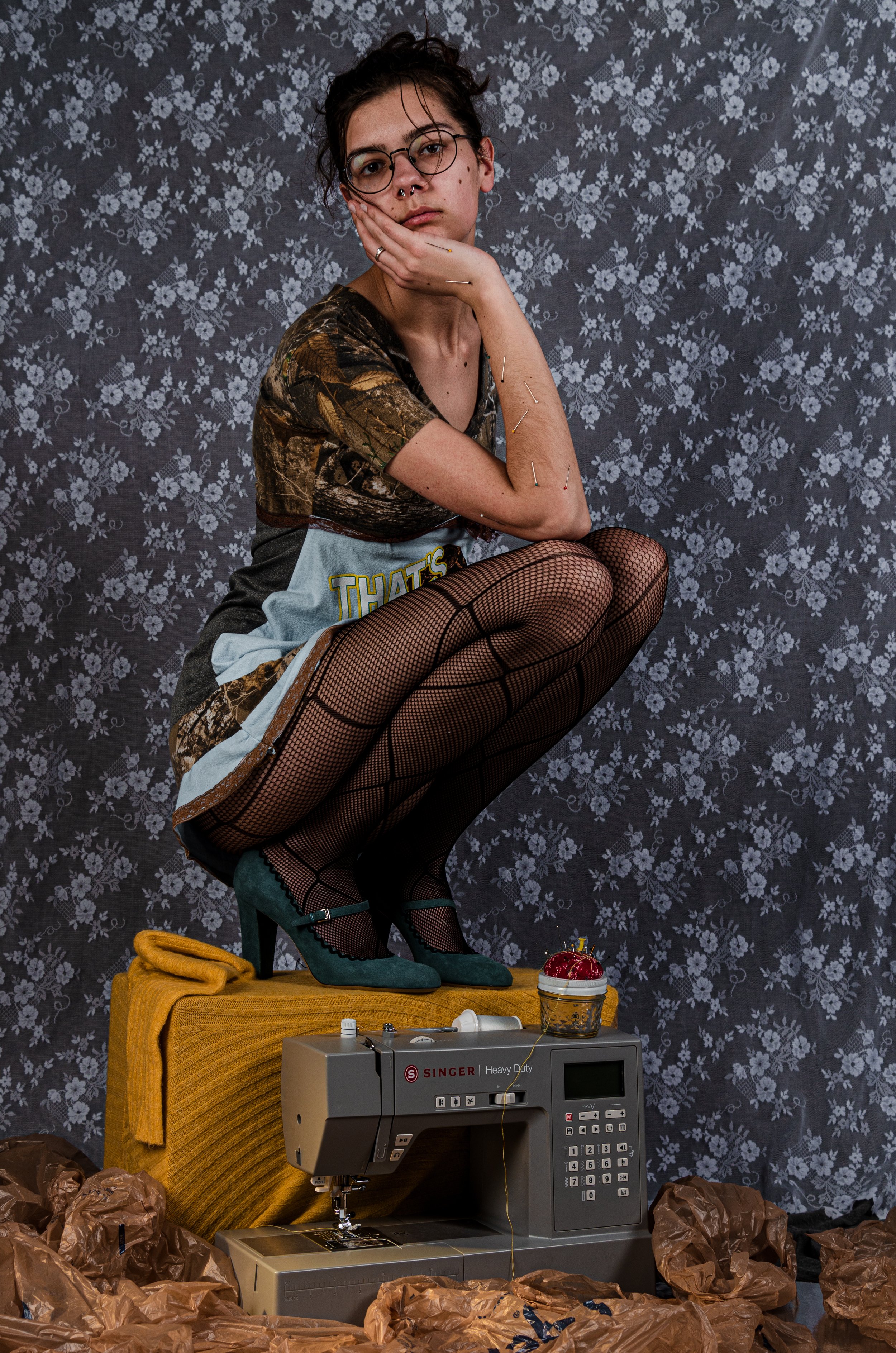 A woman crouching on a yellow upholstered surface with her chin resting on her right hand. She has short dark hair, glasses, and a septum piercing. She wears a camouflage top, fishnet stockings, and teal high heels. The background is dark floral wall