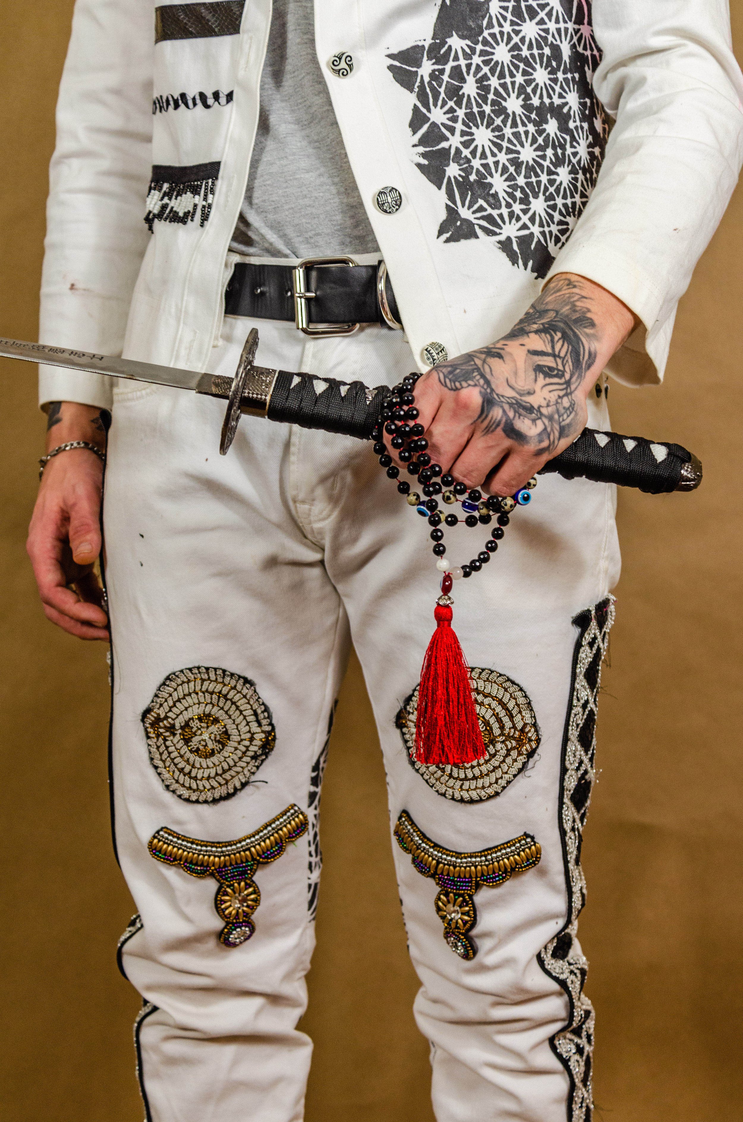 Close-up of a person's lower torso and hand holding a sword and prayer beads. The individual is wearing white embroidered pants with beaded designs and a white jacket with black and white patterns. The person also has a tattooed hand, wearing a brace