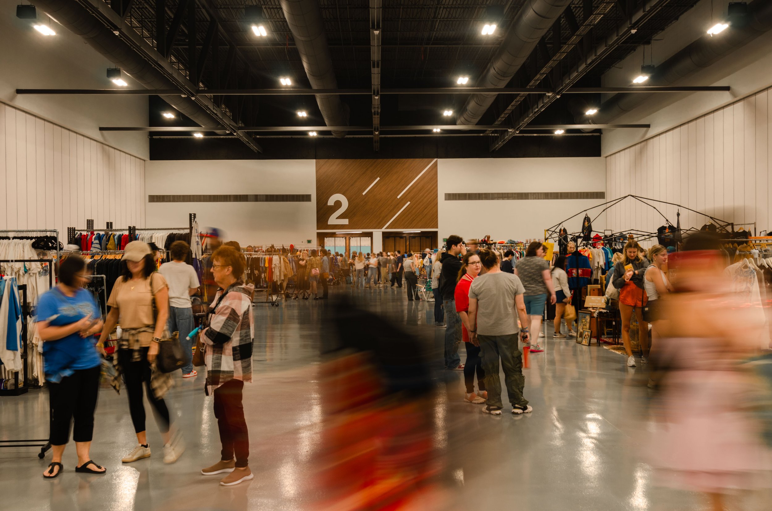 People shopping inside a large indoor marketplace with clothing racks and booths, and a large sign with the number 2 in the background.