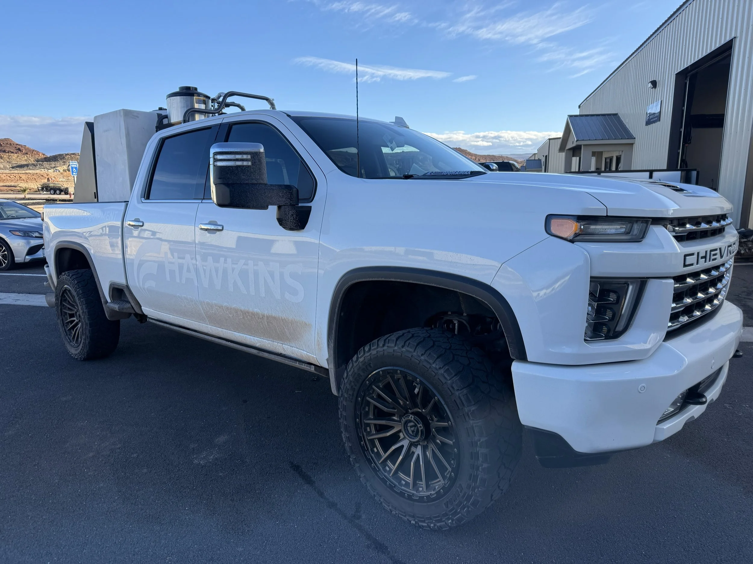White Chevrolet pickup truck with large off-road tires parked outside a building, with a mountainous landscape in the background.