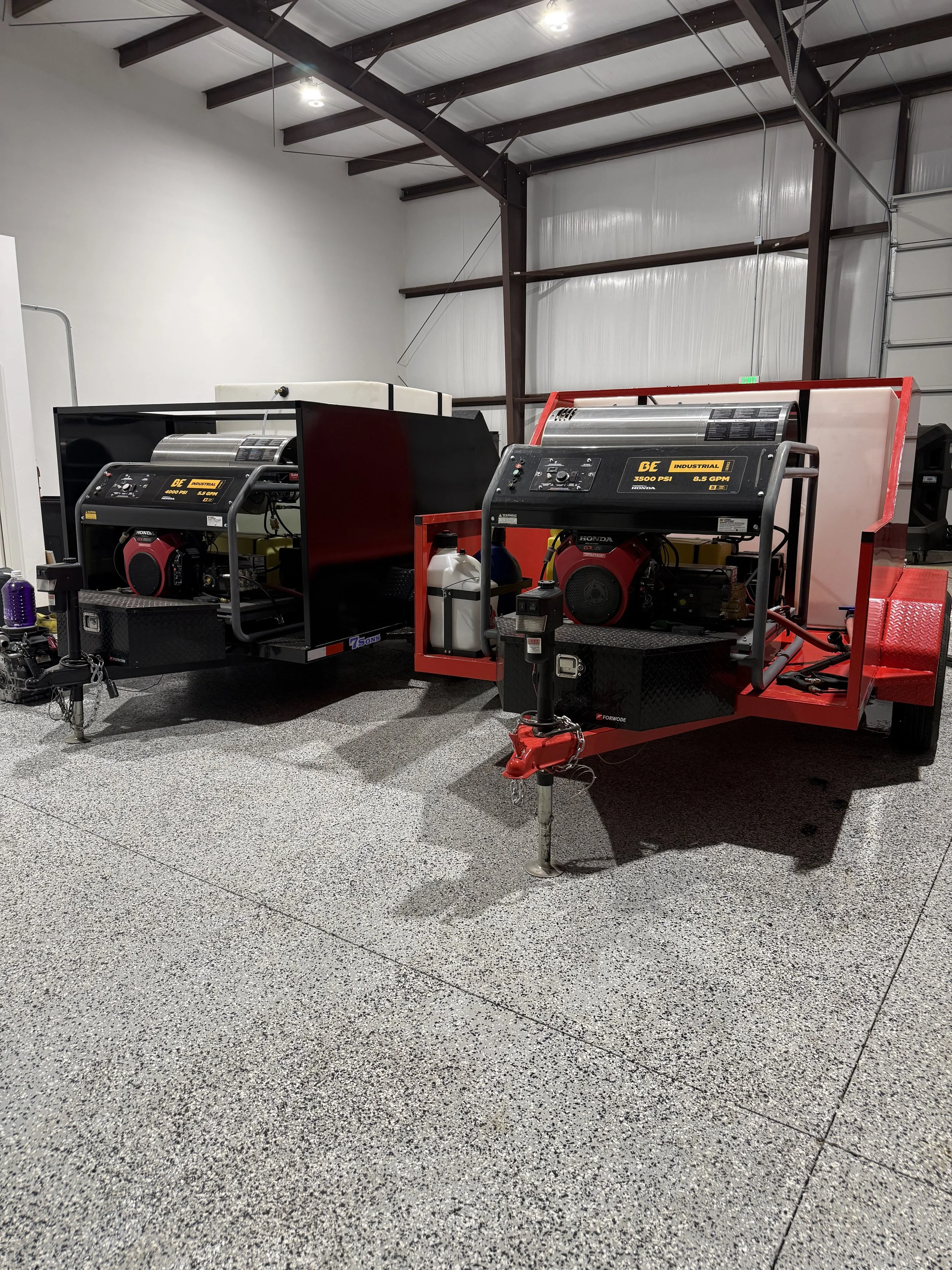 Two portable industrial hot water pressure washers on trailers inside a warehouse, with metal beams and garage doors visible in the background.