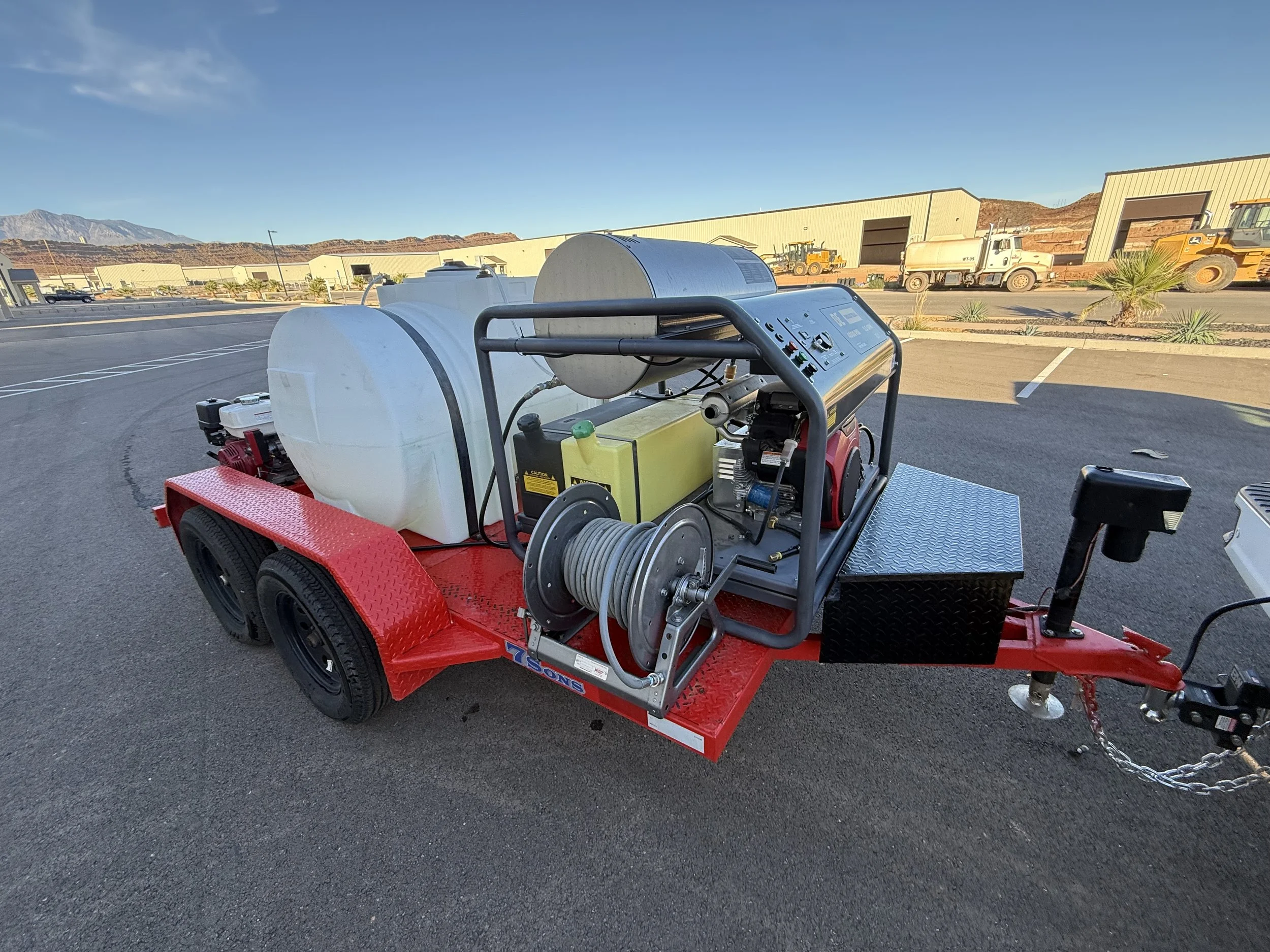 A mobile water tank and pressure washing equipment on a red trailer in an industrial lot with trucks and buildings in the background.