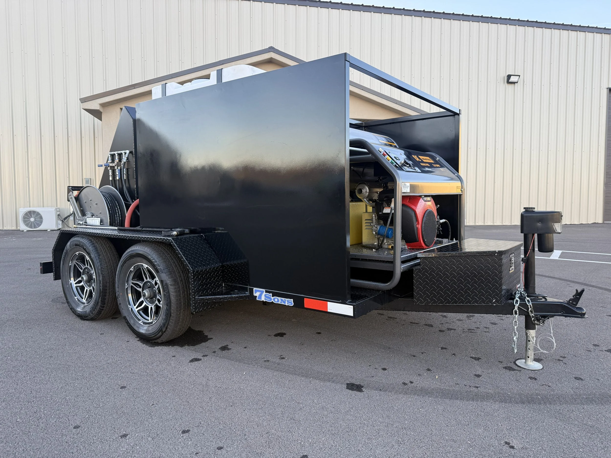 Black mobile pressure washer trailer with dual axles, hoses, and equipment, parked on asphalt in front of a beige building.