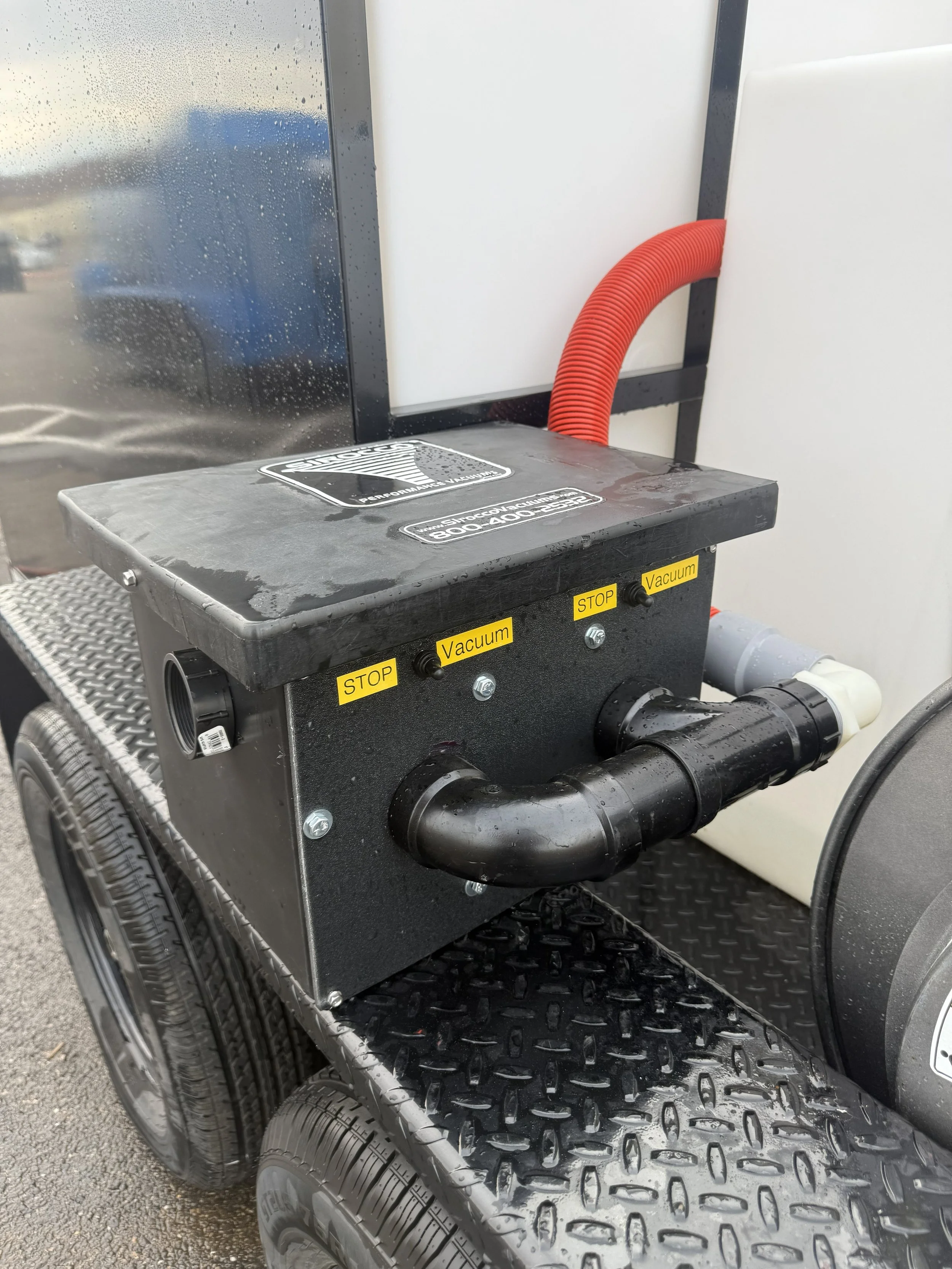 A mobile vacuum system with black metal body, labeled switches for 'STOP' and 'Vacuum', mounted on a wheeled platform with textured metal surface, and red corrugated hose attached.