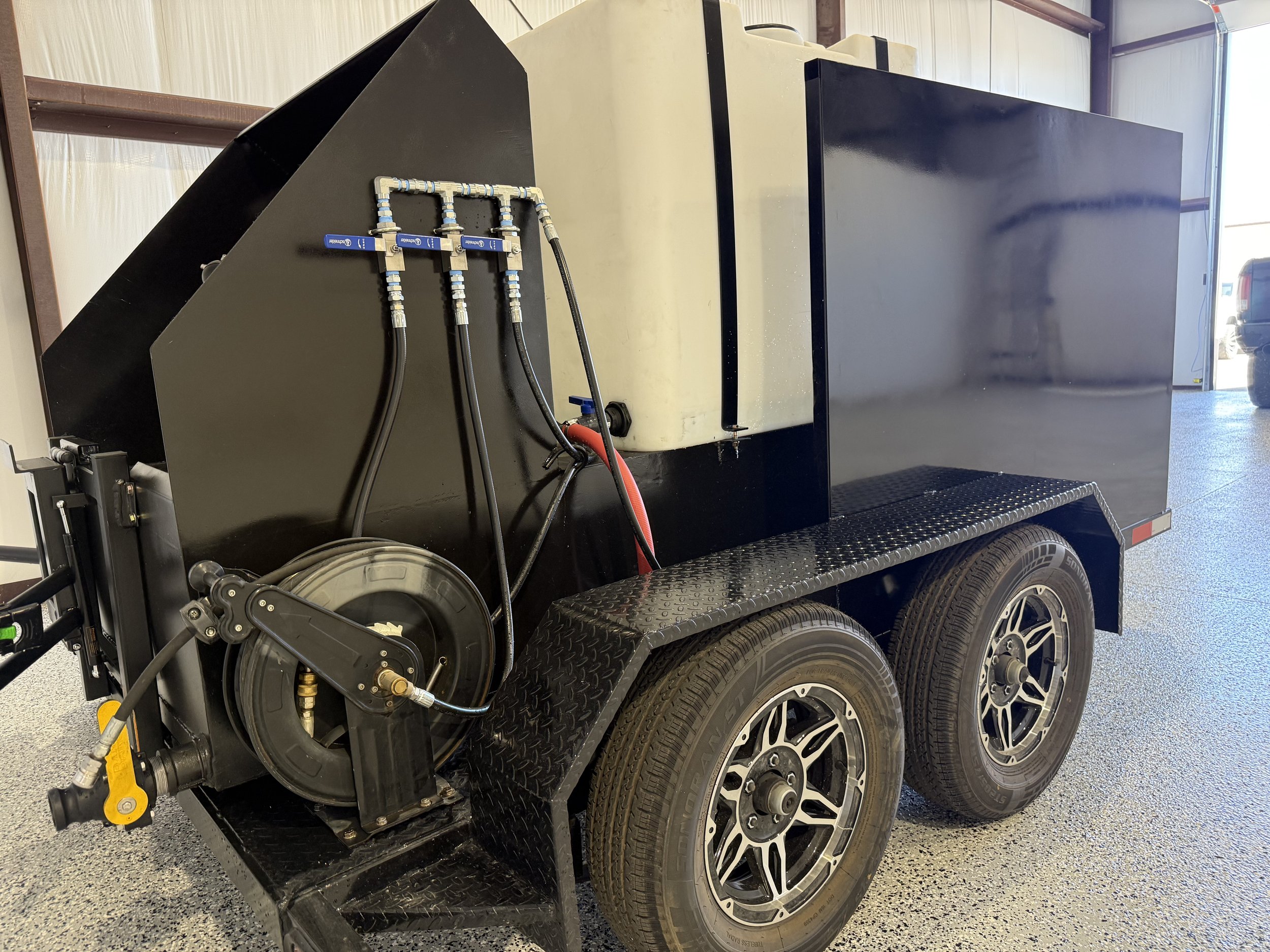 A black utility trailer with dual wheels, containing a large white water tank and equipment with hoses and valves attached to the side.