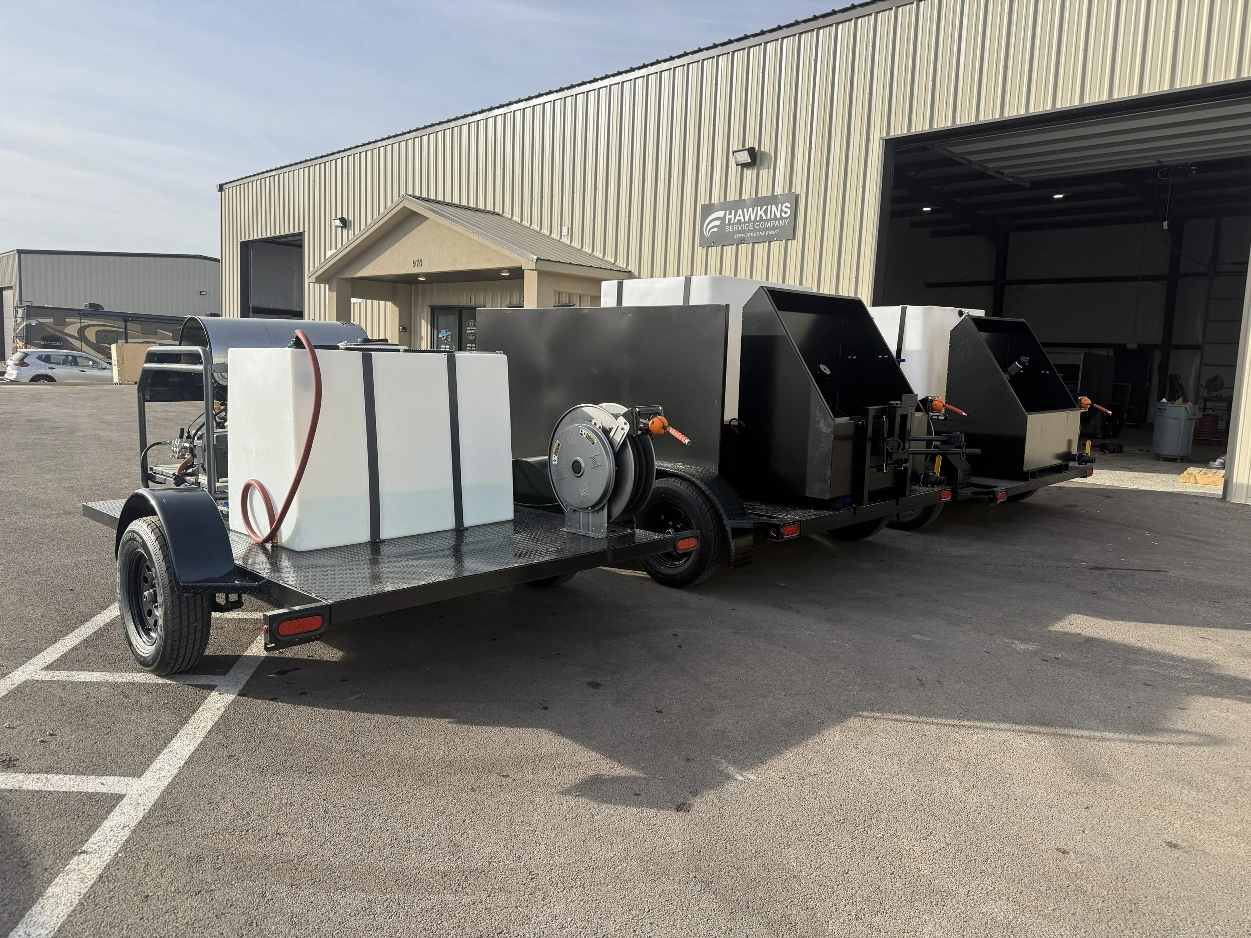 A black utility trailer with equipment, including a white tank and hoses, parked outside a warehouse with a sign that says 'Hawkins Service Company'.