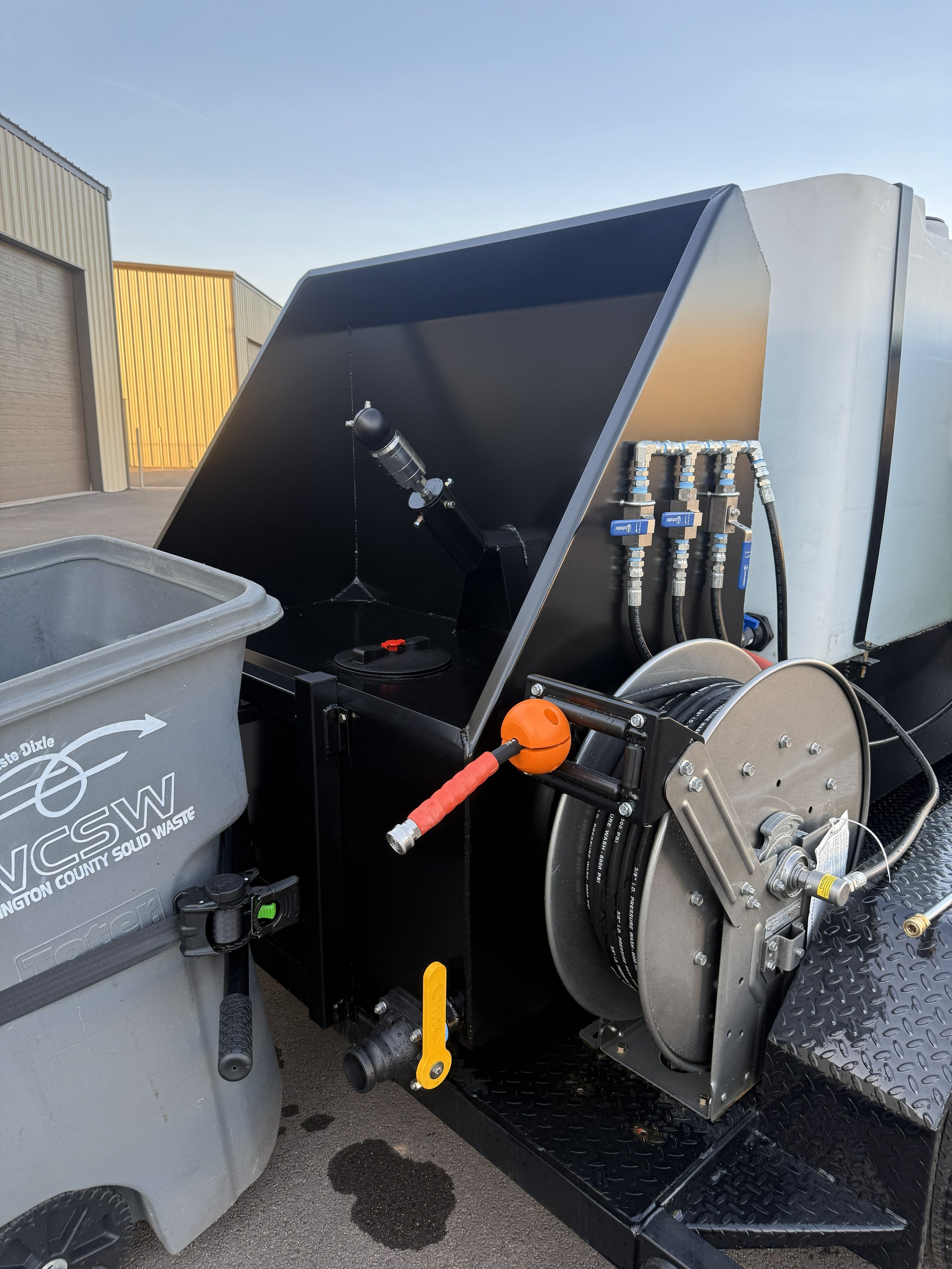 A portable wastewater cleaning or hydro jetting machine connected to a gray trash bin labeled 'WCSW Washington County Solid Waste' with a black hose on a black metal trailer.